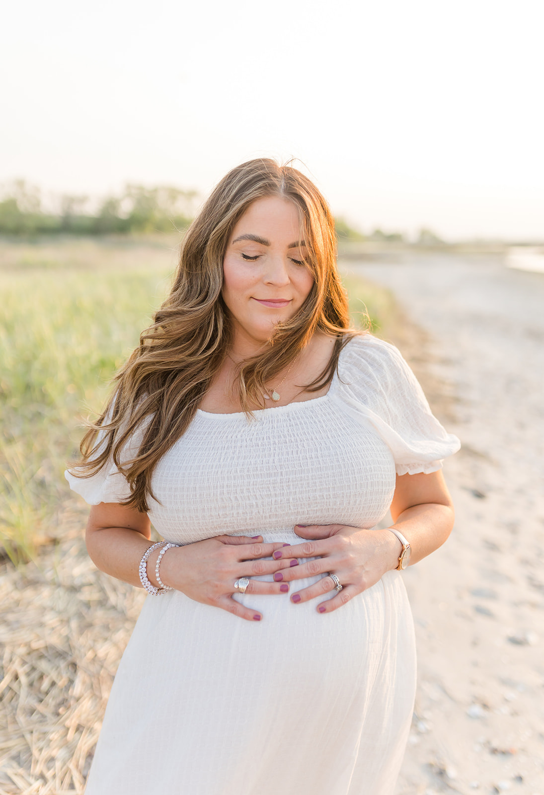 A mom to be rests her hands on the bump in a white maternity gown on a beach at sunset