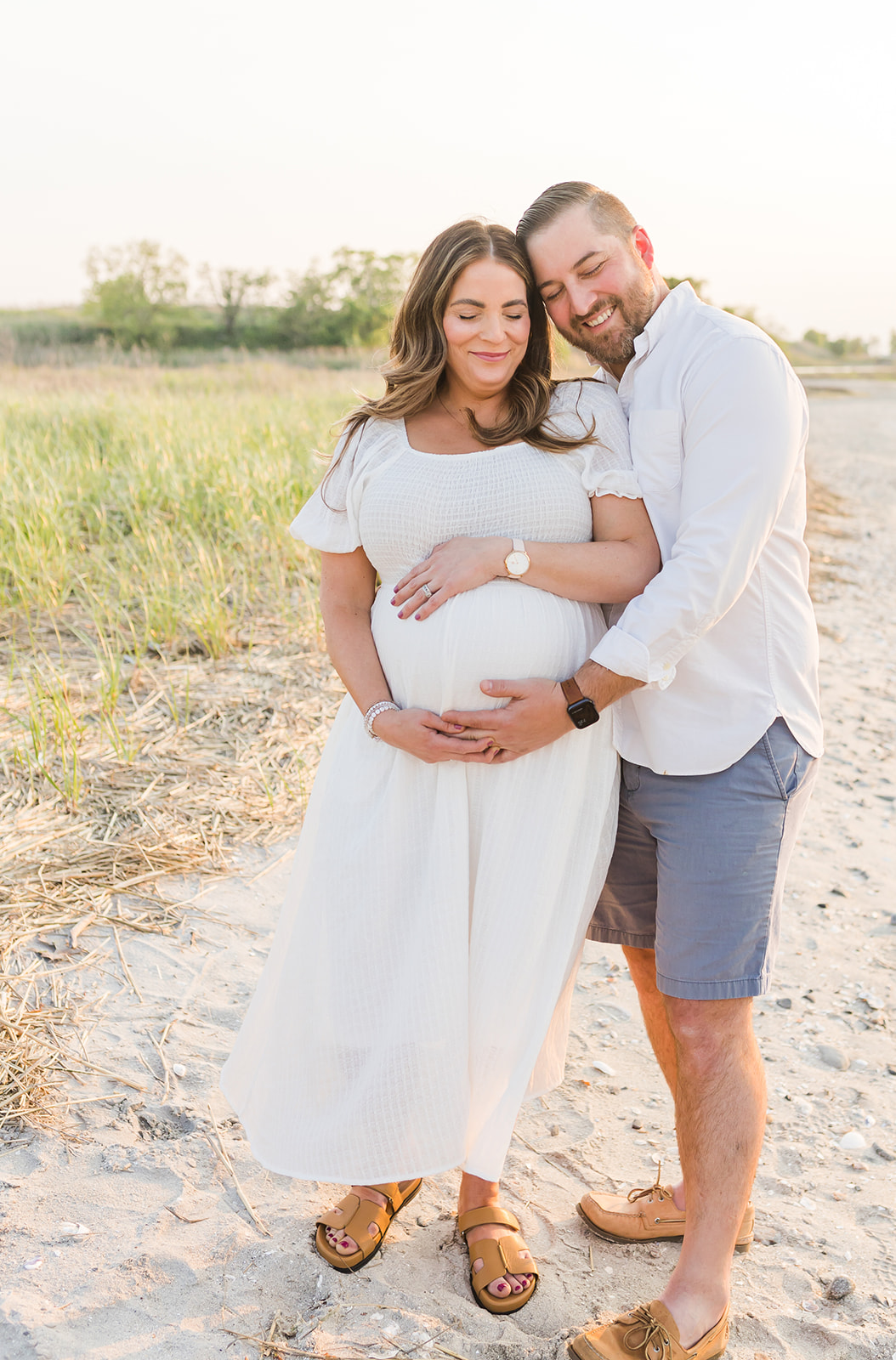 A happy dad to be holds the bump of his pregnant wife while standing on a beach at sunset before getting a 3D Ultrasound In Connecticut