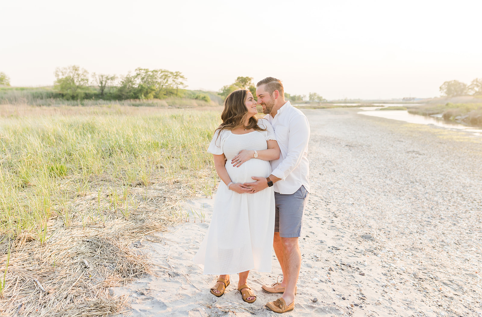 A mom to be smiles while being wrapped up by her husband in a park by a river at sunset before getting a 3D Ultrasound In Connecticut