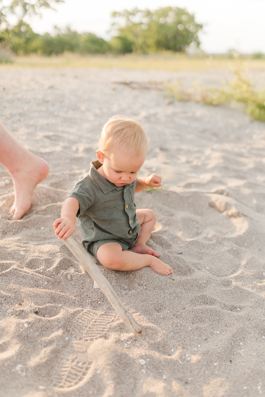 A toddler boy in a green onesie plays with a stick in the beach sand at sunset before visiting a Child Therapist Fairfield CT