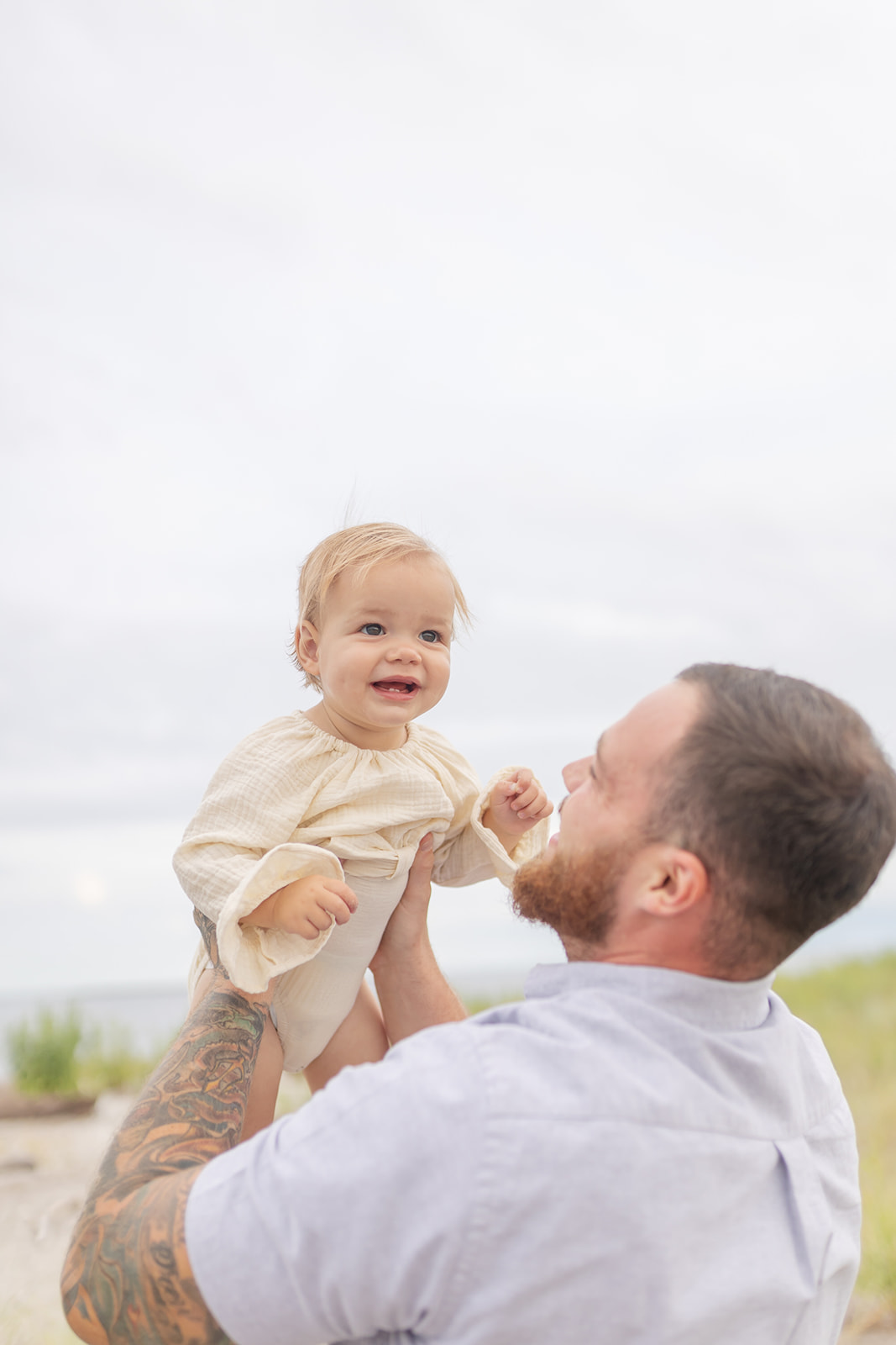 A father in a blue shirt lifts his toddler daughter above his head on a beach