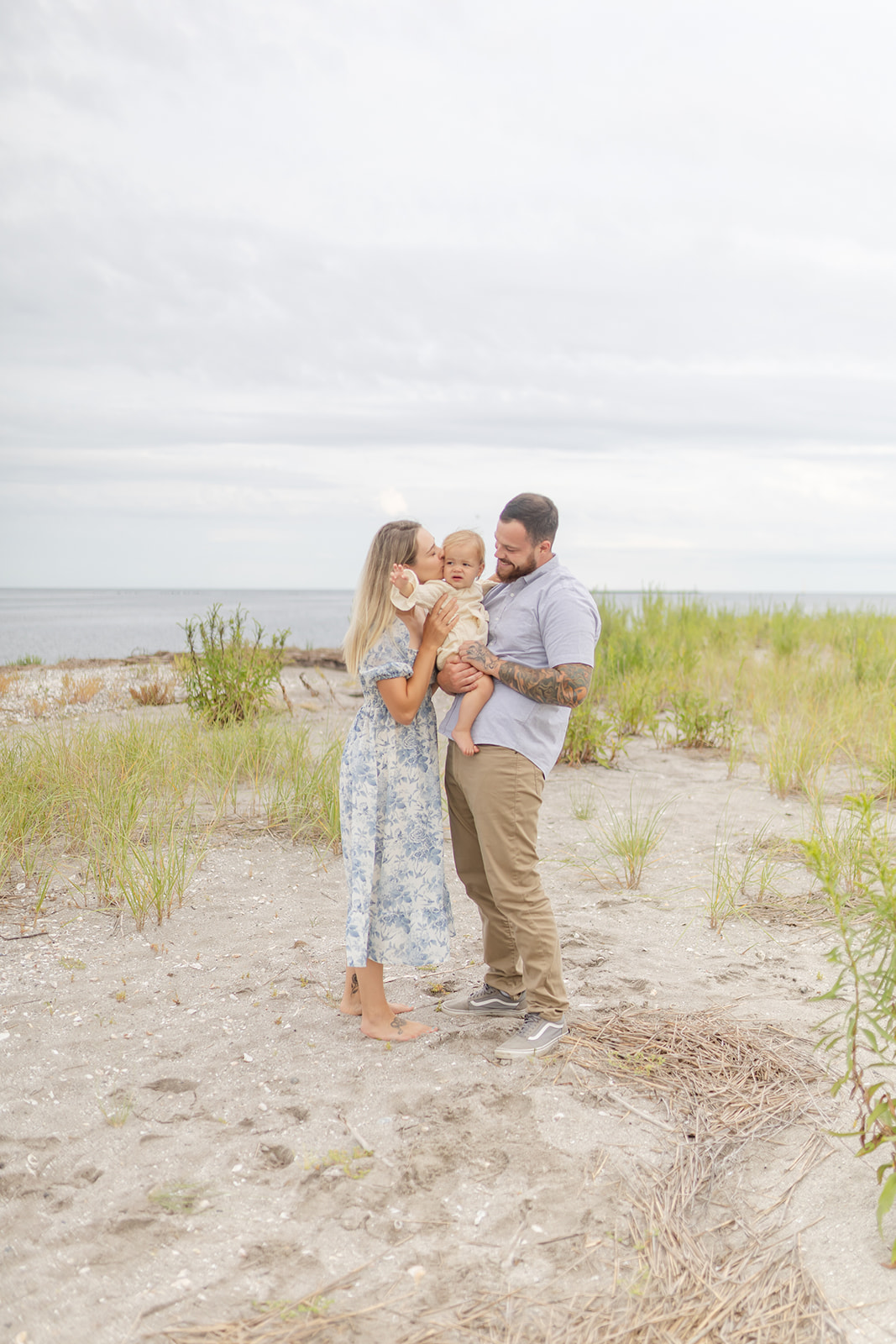 Parents in blue kiss and play with their toddler daughter in dad's arms on a beach after visiting Groove Westport