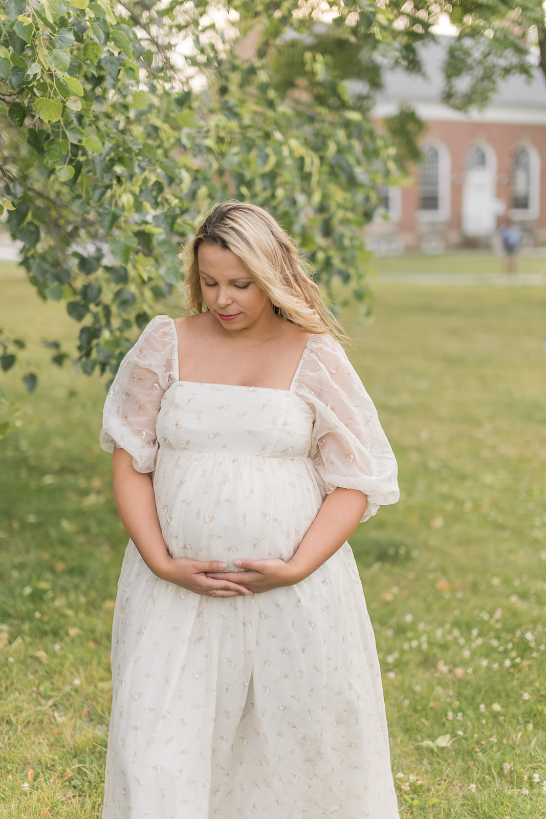 A mom to be in a white maternity gown stands under a tree in a park holding her bump