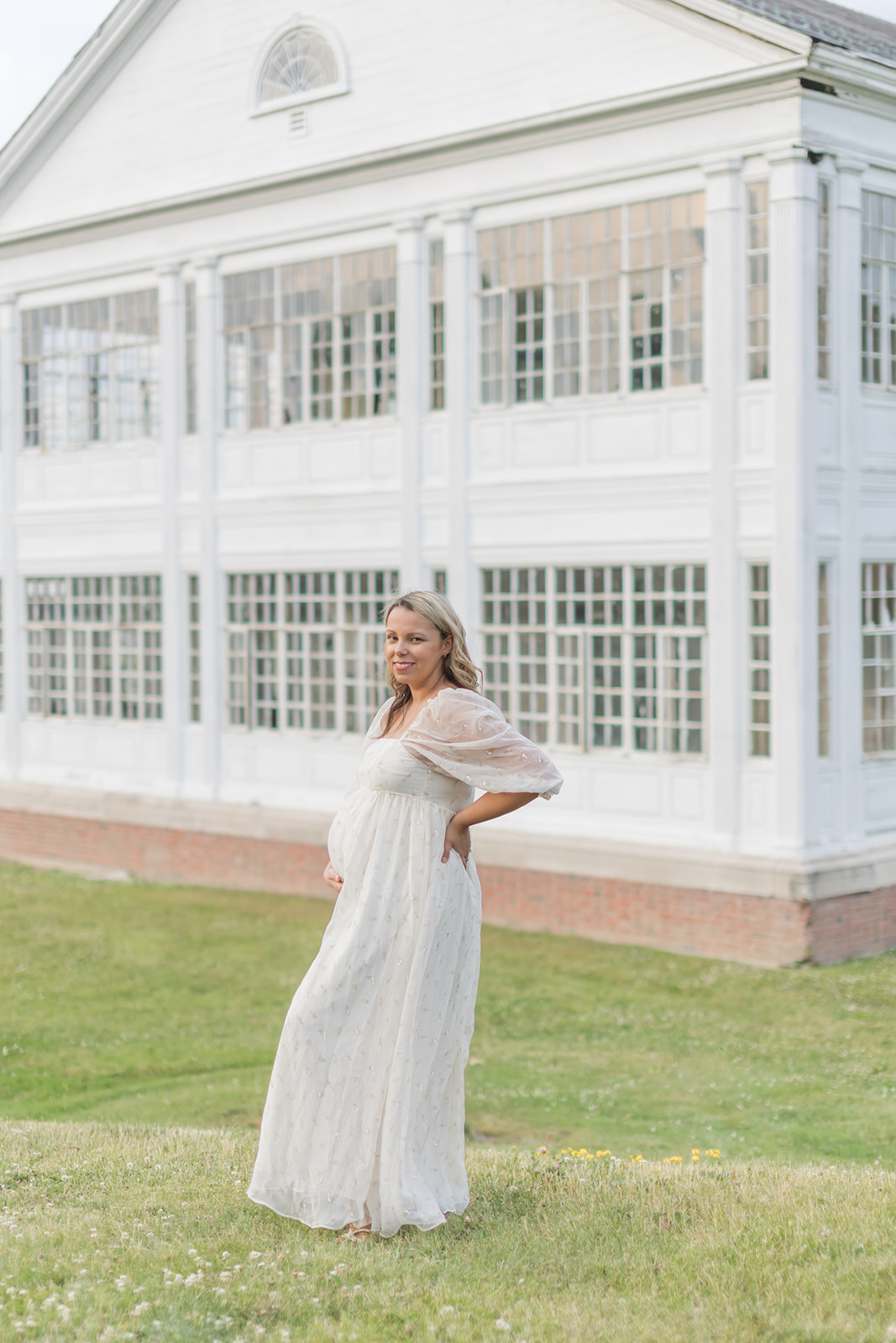 A mother to be stands in a white lace maternity gown in a field in front of a colonial building holding her bump before a Home Birth CT