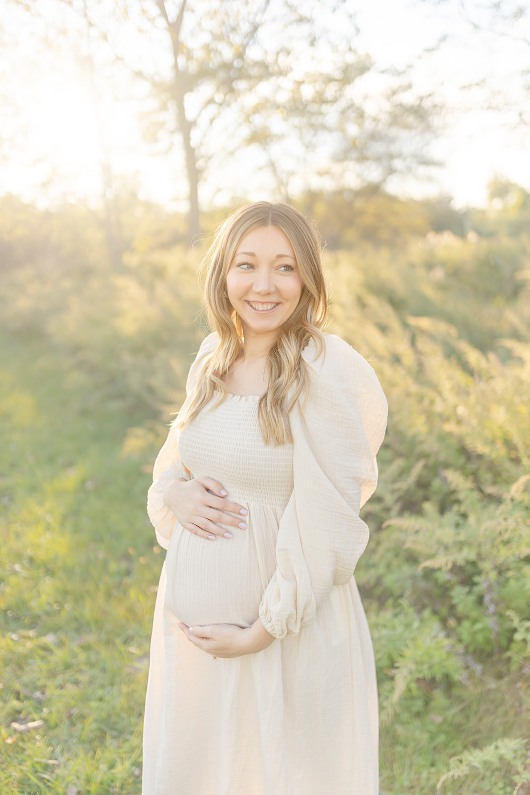 A mom to be smiles over her shoulder while holding her bump in a park at sunset