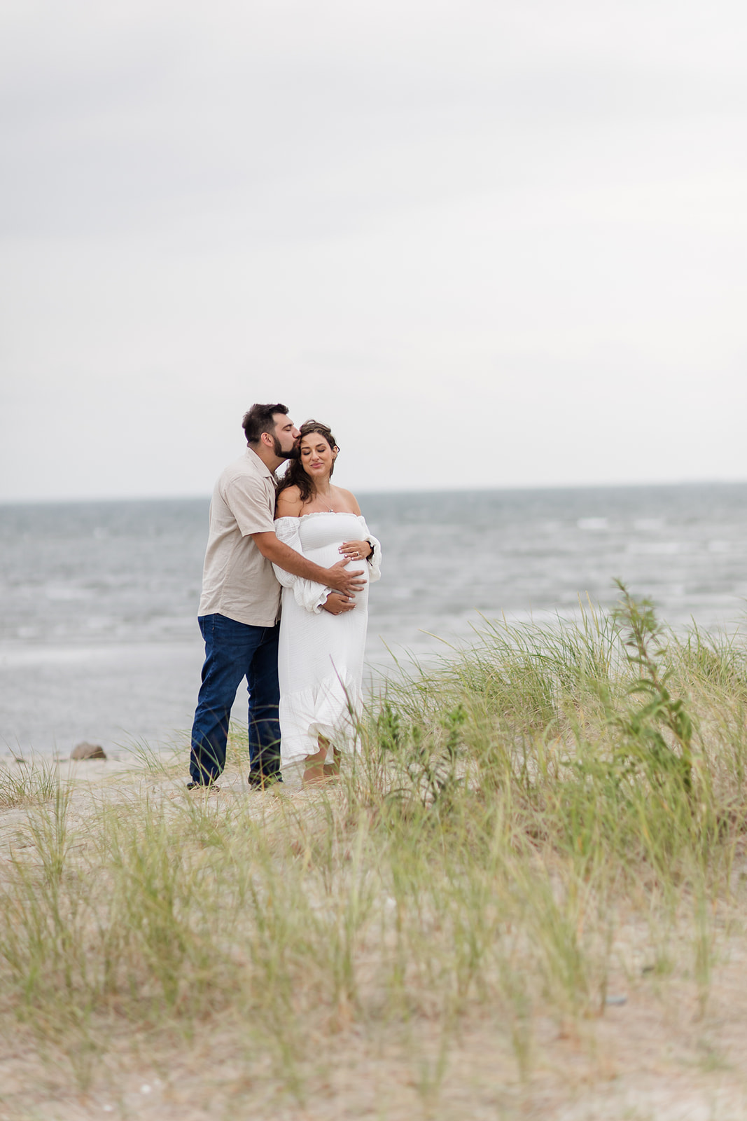 A mom to be stands on a windy beach while being kissed by her husband and holding the bump before meeting with their Postpartum Doula CT
