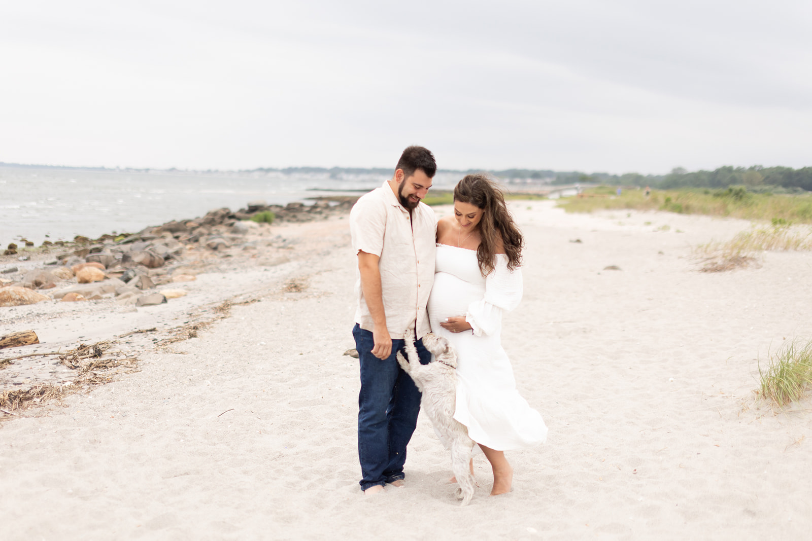 A mom to be in a white dress stands with her husband as they play with their white dog on a windy beach after meeting with their Postpartum Doula CT
