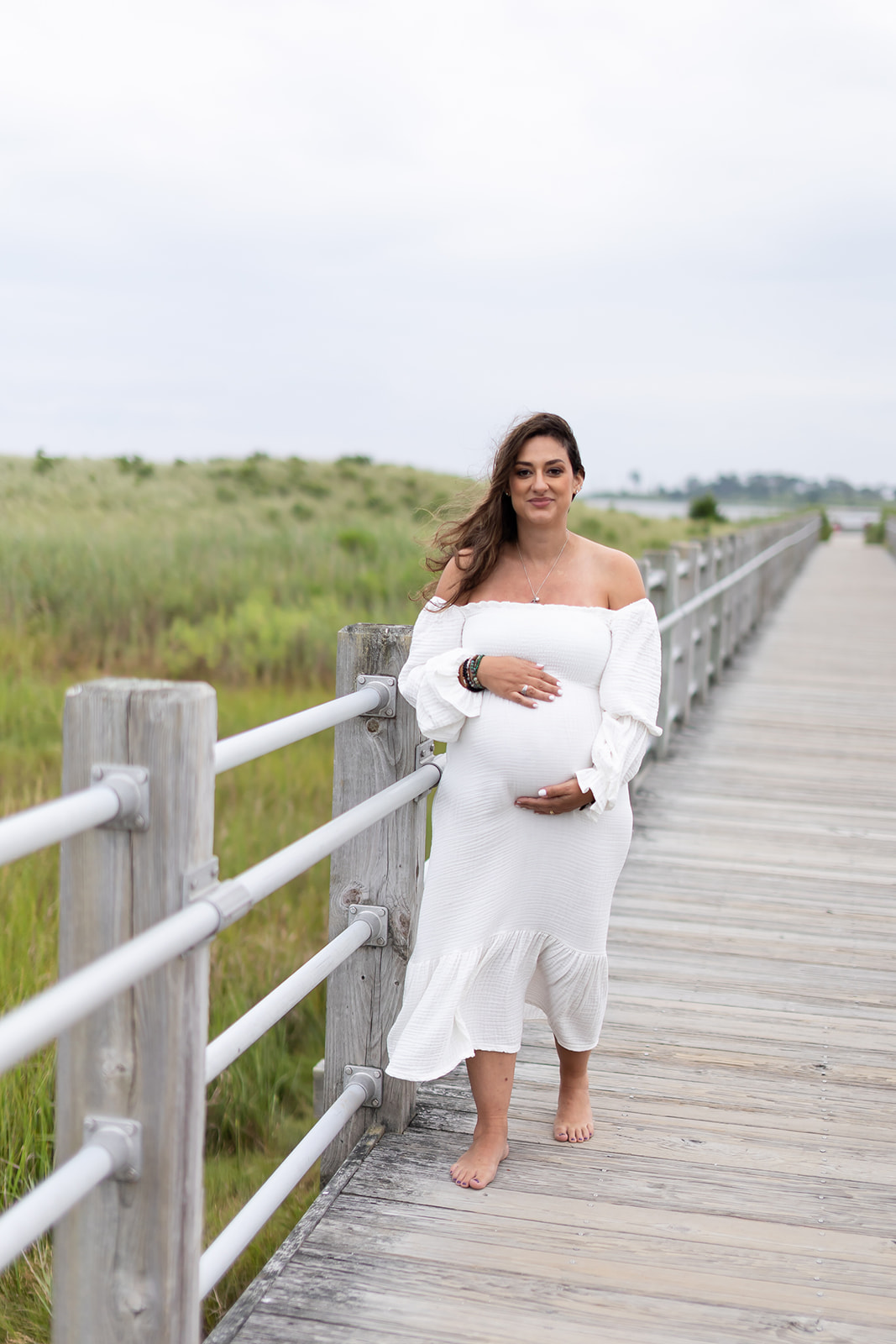 A mom to be walks barefoot on a wooden boardwalk at the beach in a white dress while holding her bump
