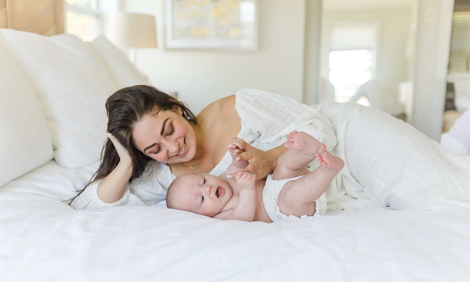 A new mom in a white dress lays on a bed playing with her infant baby after visiting Stroller Friendly Trails CT