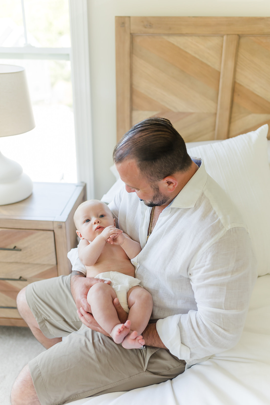 A father in a white shirt sits on the edge of a bed looking down at his newborn baby in his arms before walking on Stroller Friendly Trails CT