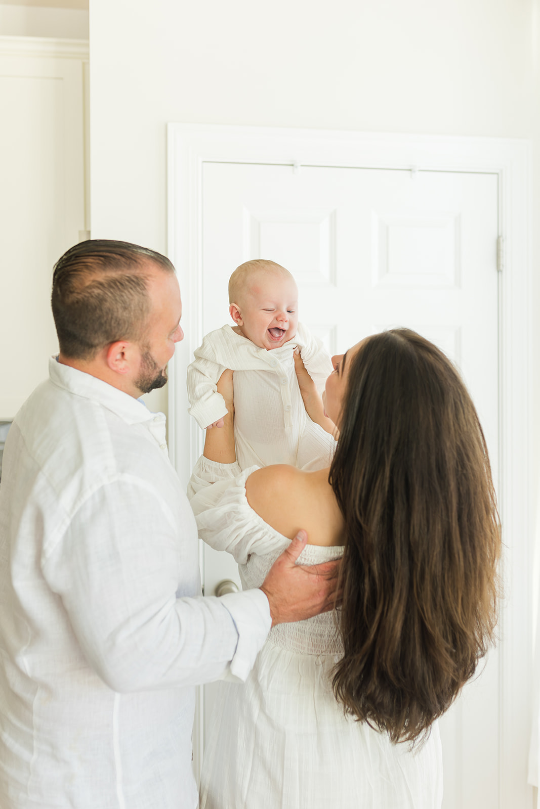 A mom and dad play with their smiling newborn baby in a whtie nursery