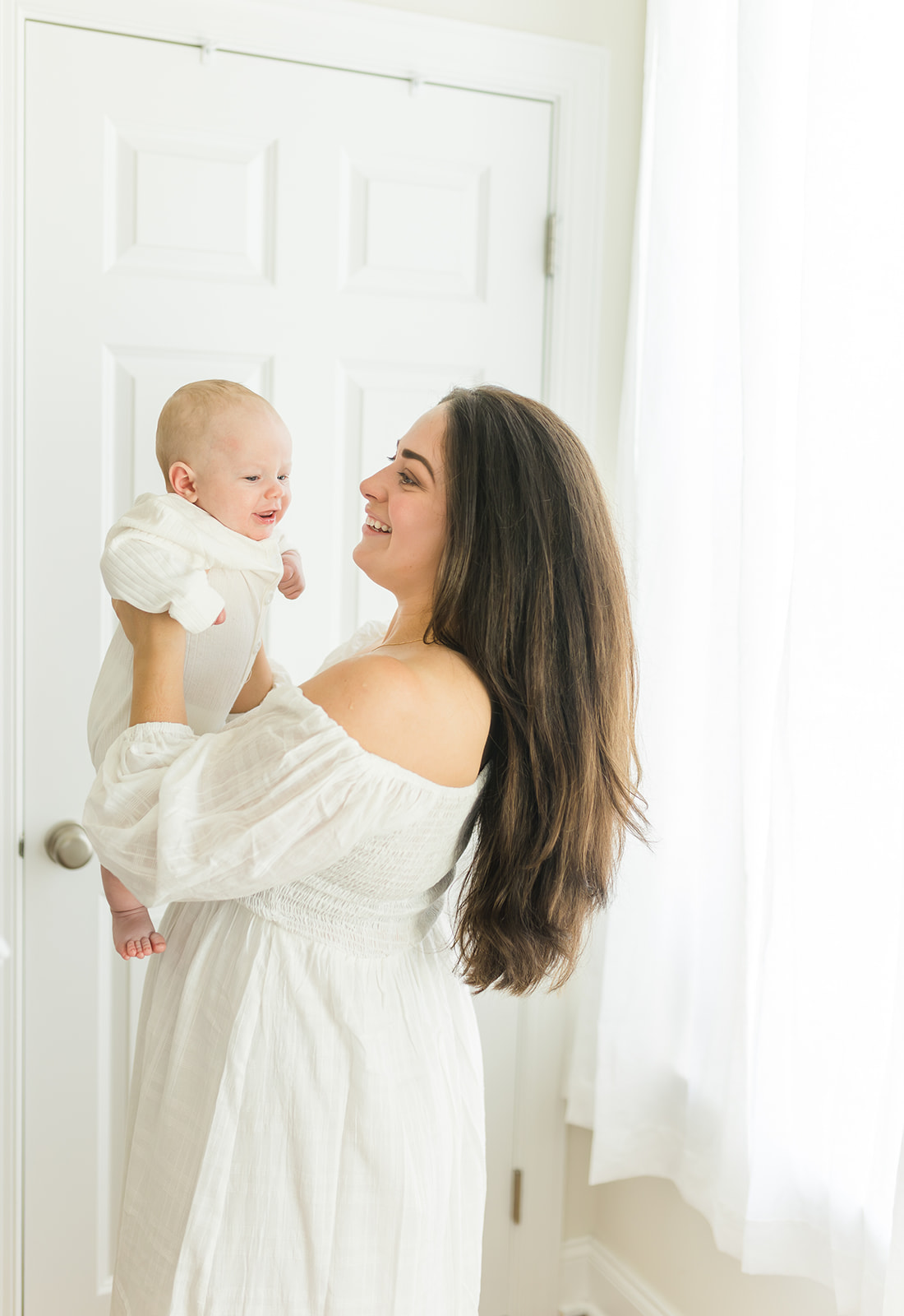 A mother in a white dress lists her laughing newborn baby by a window after visiting Tiny Miracles