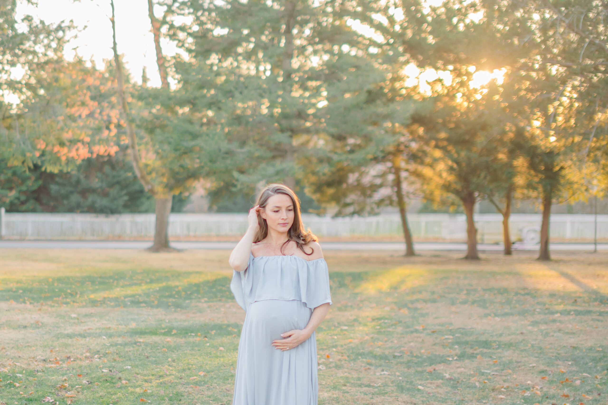 pregnant woman in a blue off the shoulder dress standing in front of a white picket fence with sun bursting through the trees