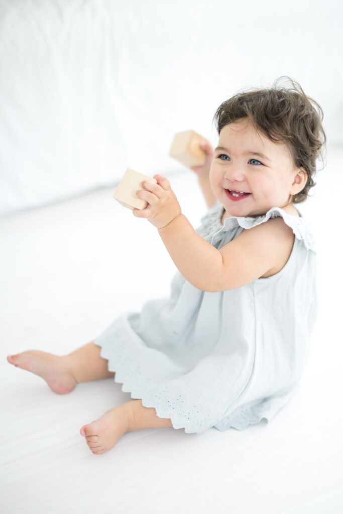 little girl with brown hair in a blue dress holding some blocks and celebrating turning one years old