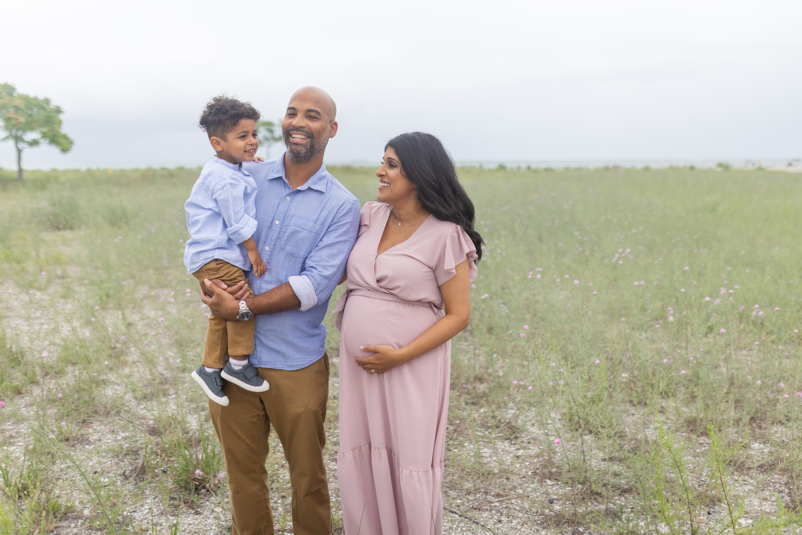 A father in a blue shirt and brown pants stands in a field holding his toddler son in matching clothes while mom holds her pregnant bump in a pink maternity gown Trumbull Pediatricians