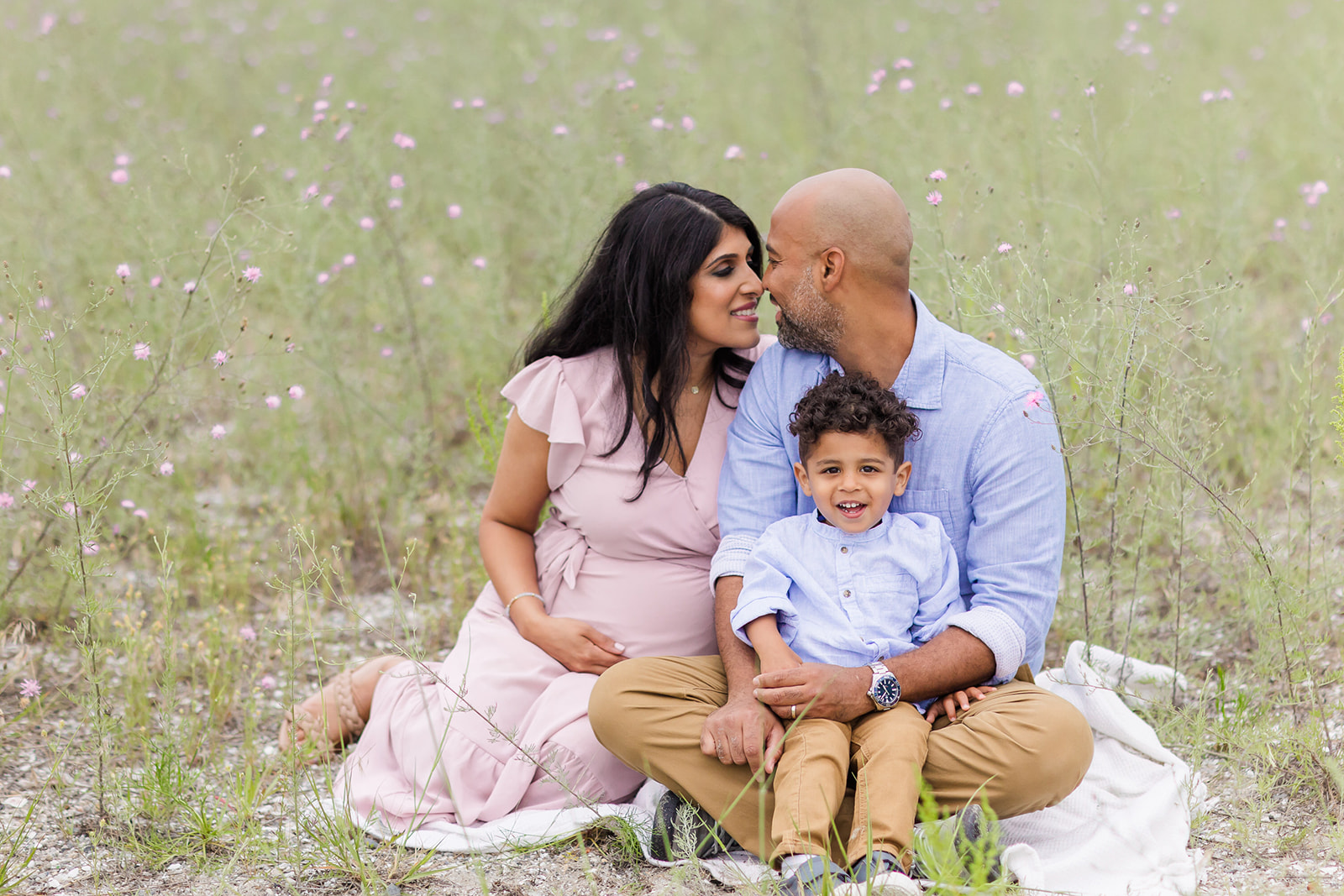 A pregnant mom and dad sit on a picnic blanket in a field of wildflowers with their toddler son in dad's lap after visiting Trumbull Pediatricians