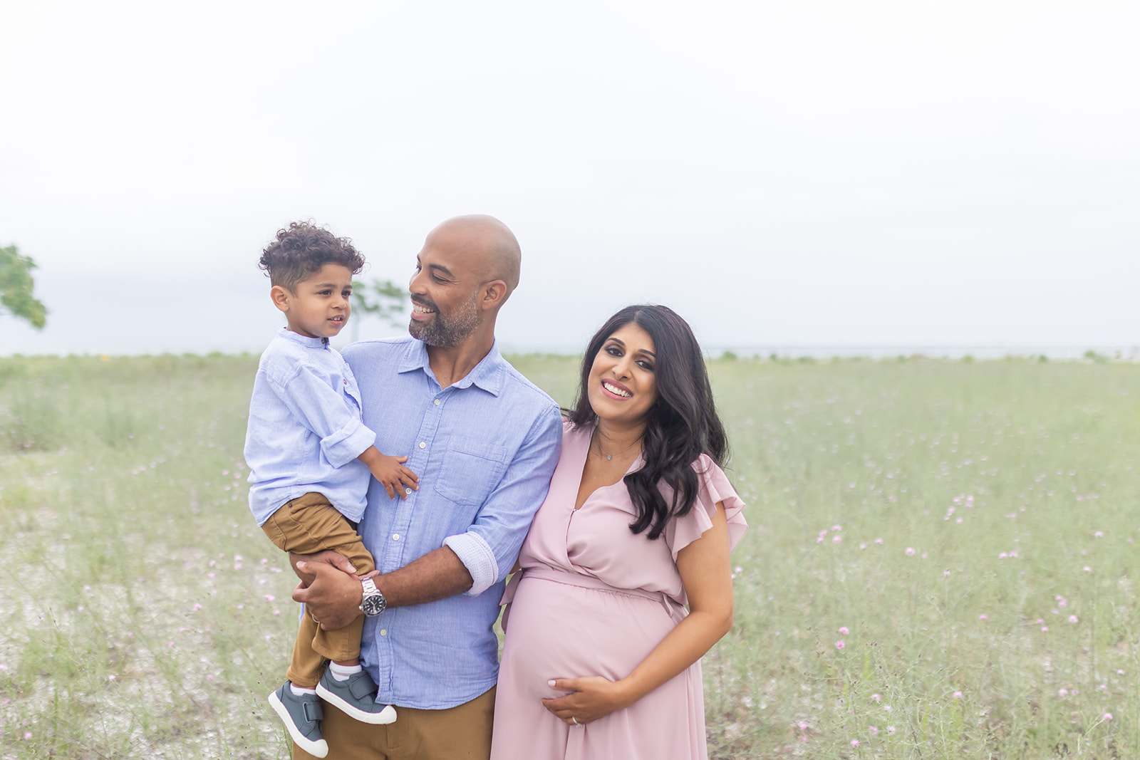 A mom to be holds her bump while leaning against her husband holding their toddler son in a field