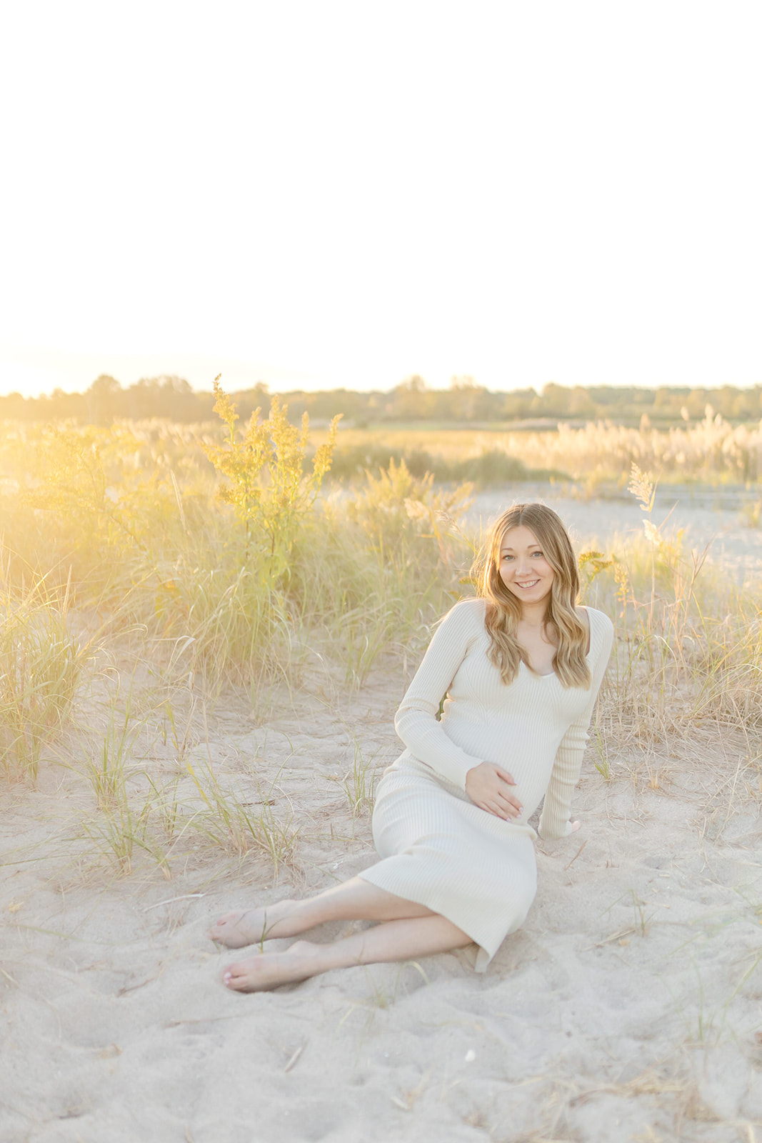 A mom to be sits in the dunes on a beach holding her bump at sunset