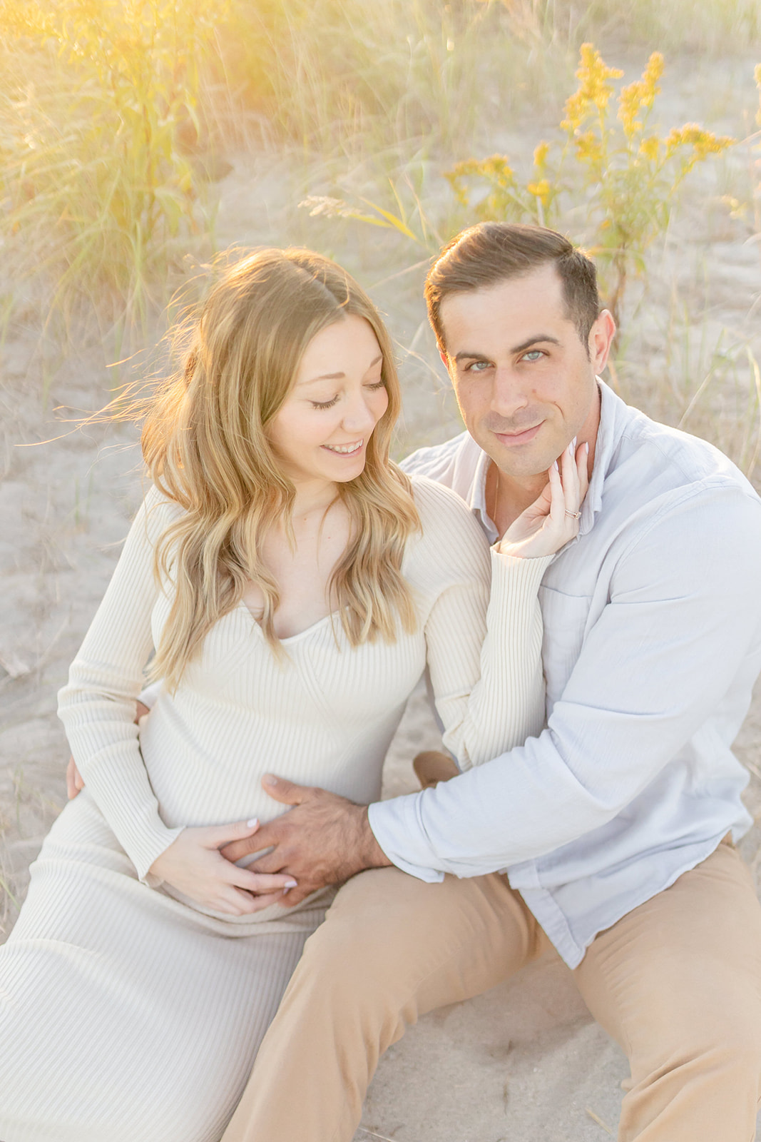 A mom to be holds her husbands chin as they sit together in the sand on a beach at sunset holding the bump after Birthing Classes CT