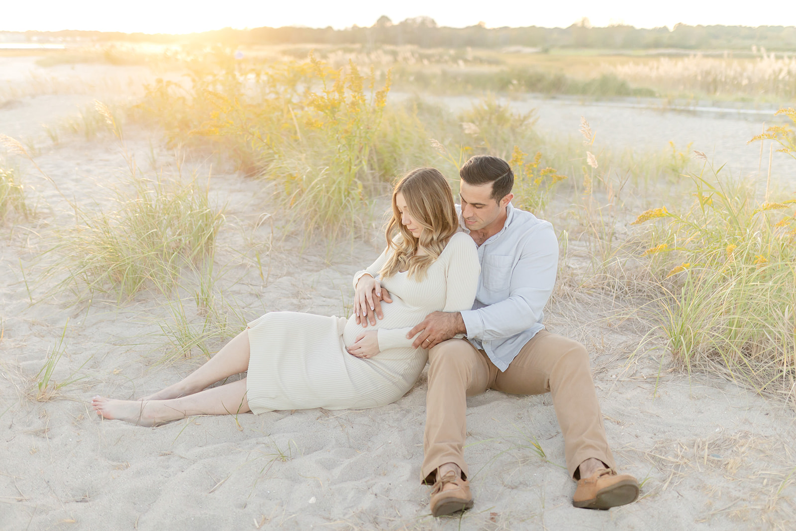 Expecting parents snuggle on a beach while holding the bump in a white dress after Birthing Classes CT