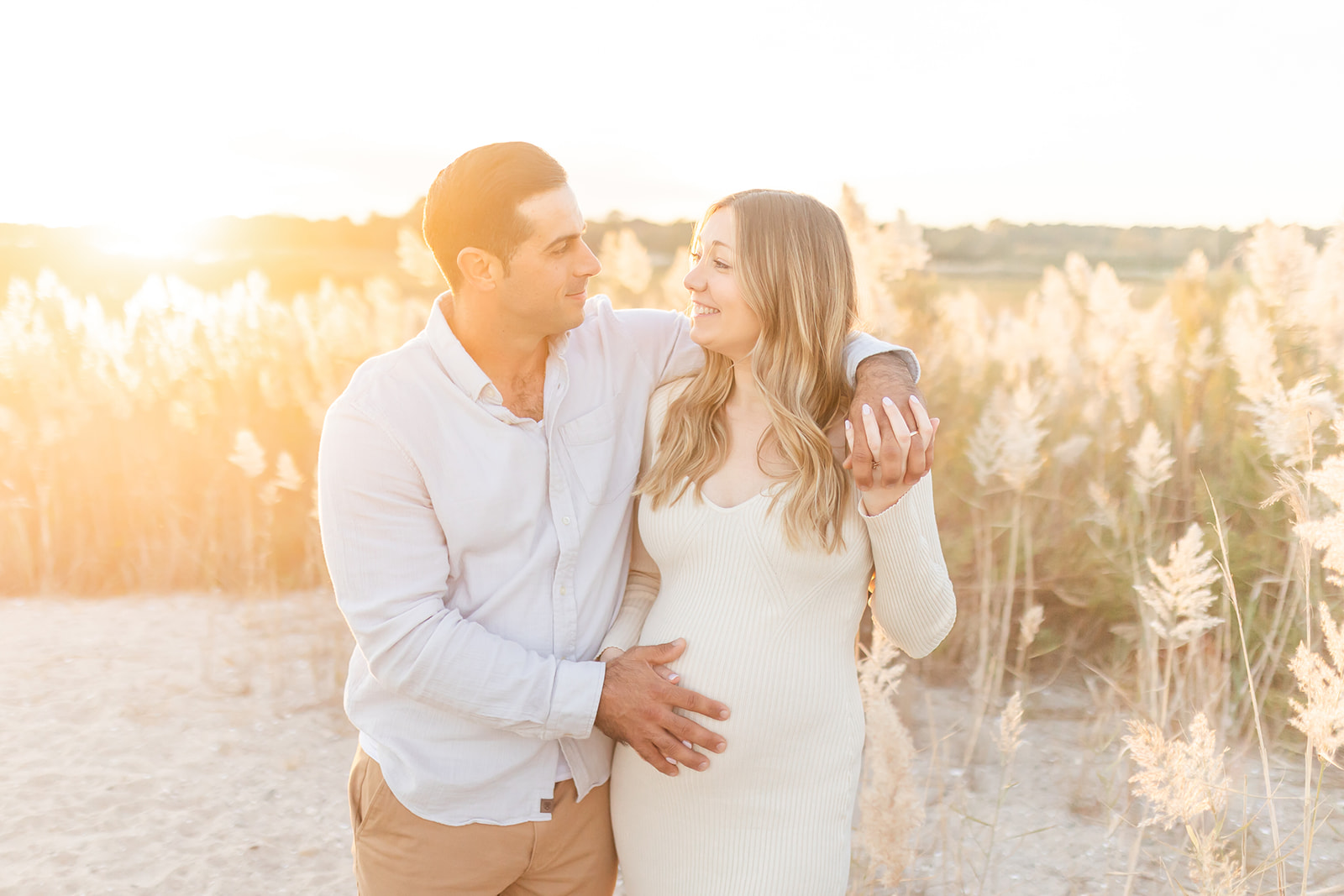 Expecting parents rest a hand on the bump while walking through a field of tall golden grasses at sunset