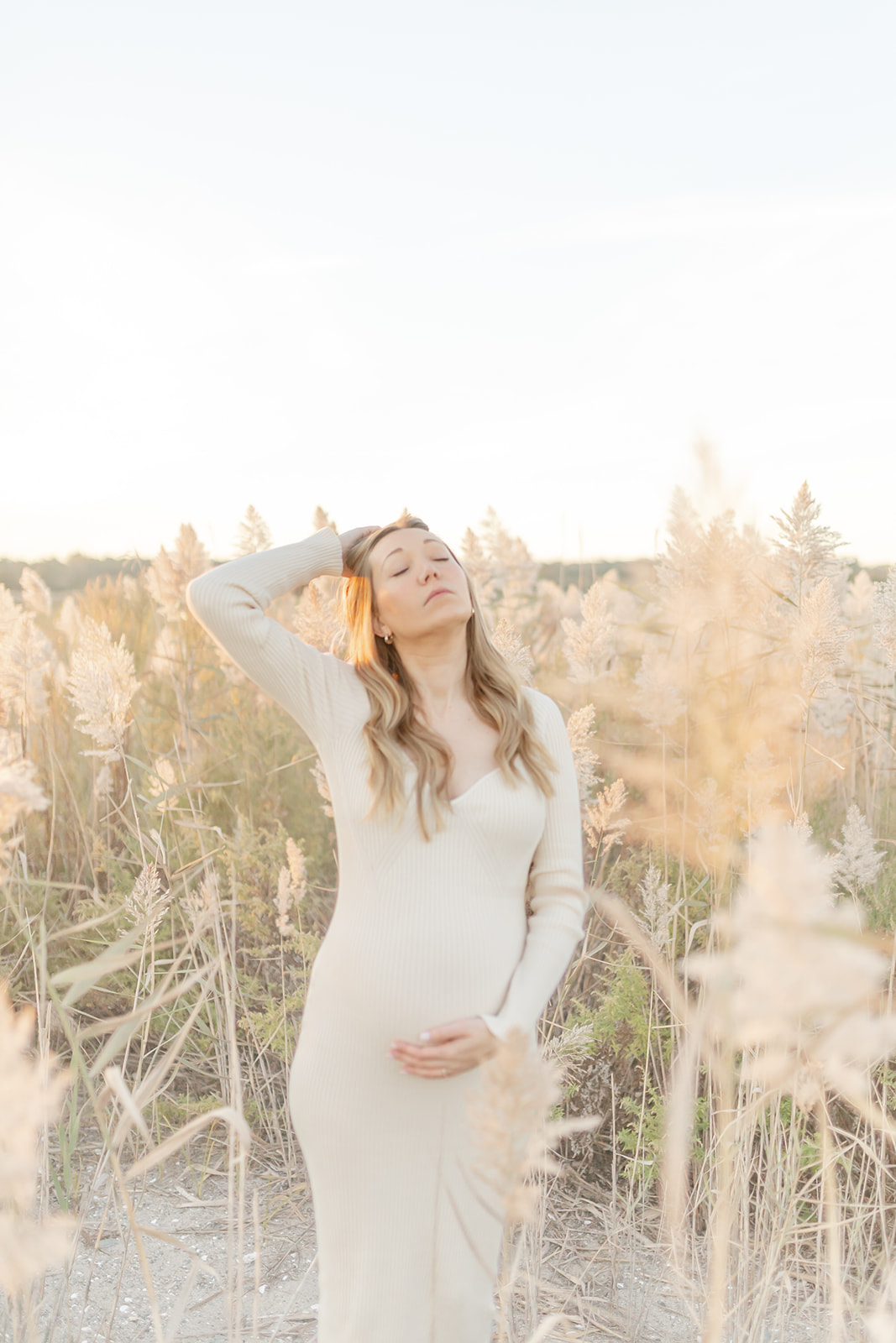 A mom to be stands in a field of tall golden grass at sunset with a hand in her hair and one on her bump before visiting Griffin Hospital labor and delivery