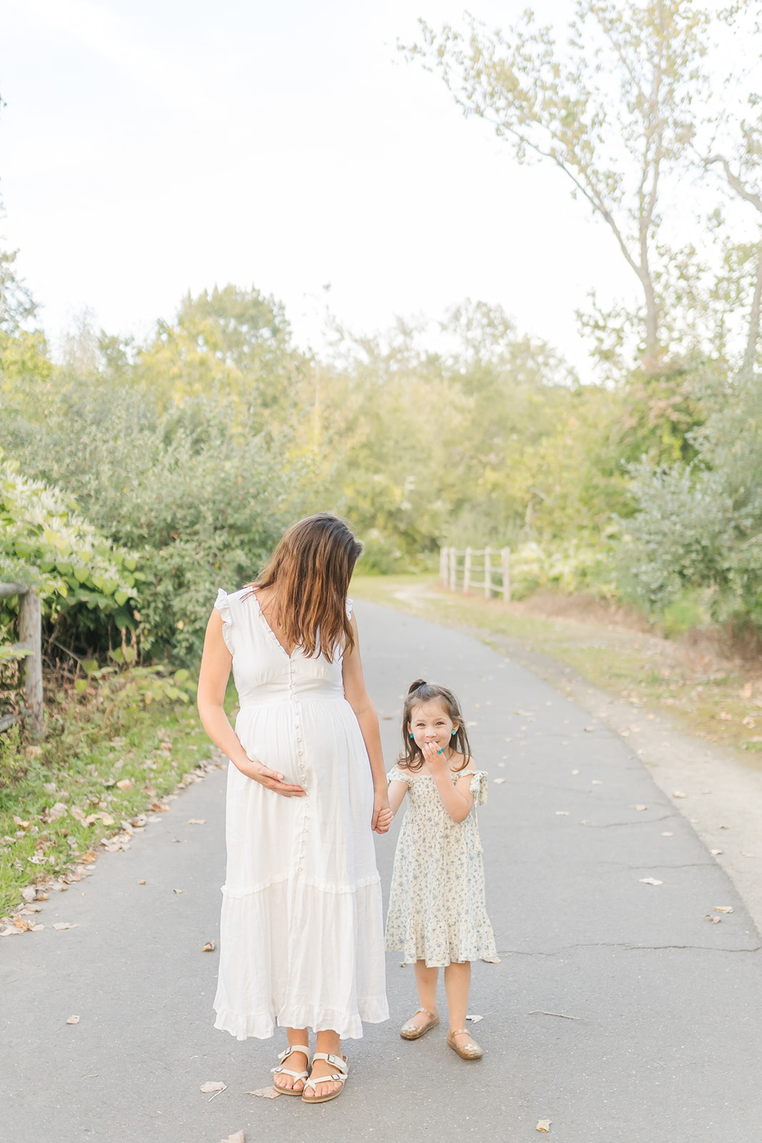 A pregnant mother in a white maternity dress walks down a park sidewalk while holding hands with her young daughter