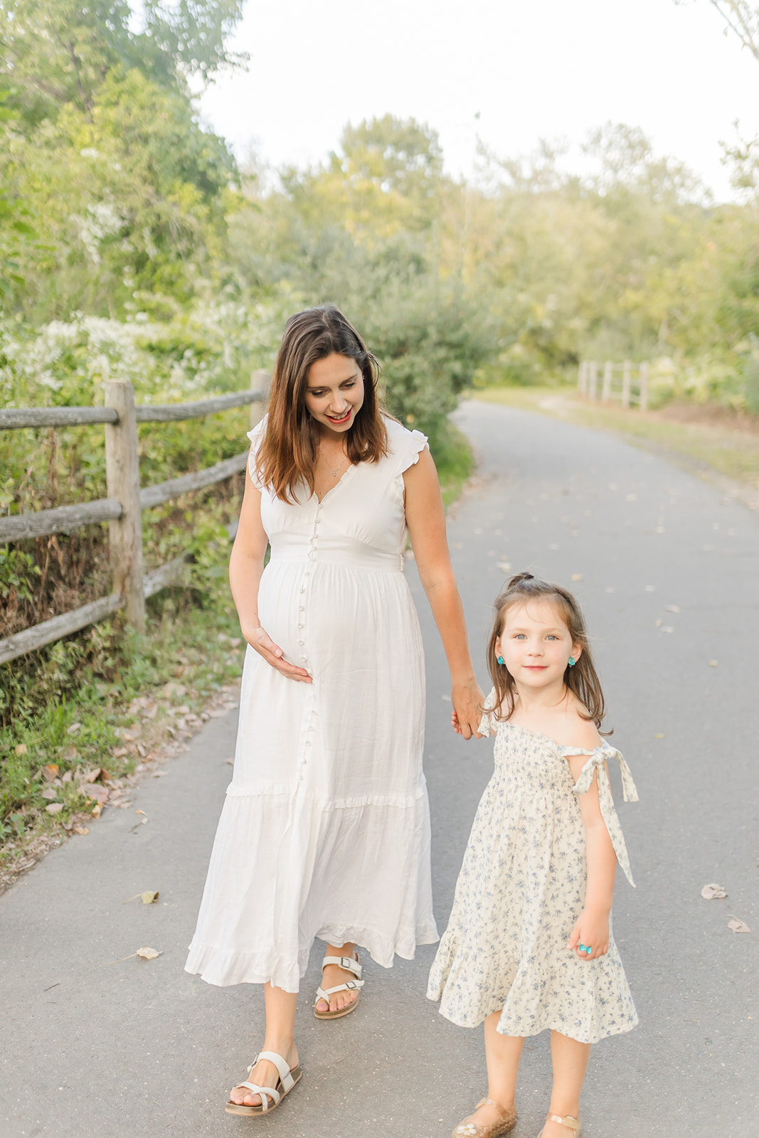 A young girl in a floral dress leads her pregnant mom by the hand through a park sidewalk during Mommy and Me Classes Fairfield CT