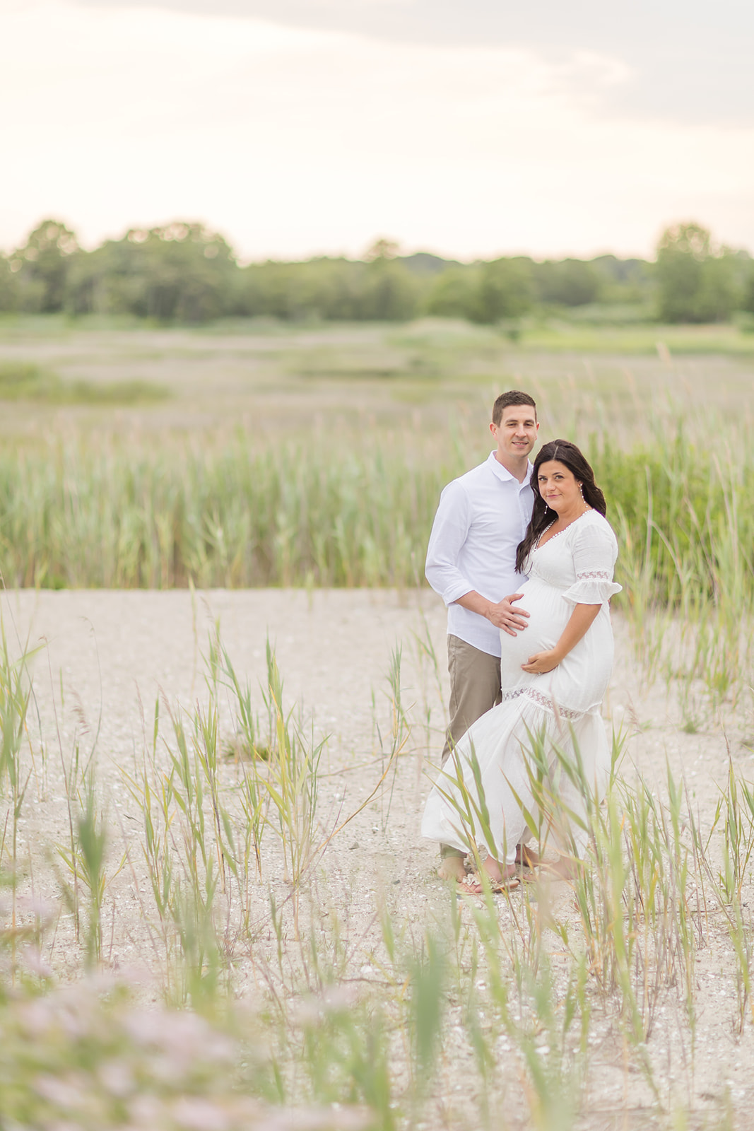 A mom to be in a white maternity dress stands on the beach surrounded by tall grasses with her husband after visiting St. Vincents Birthing Center