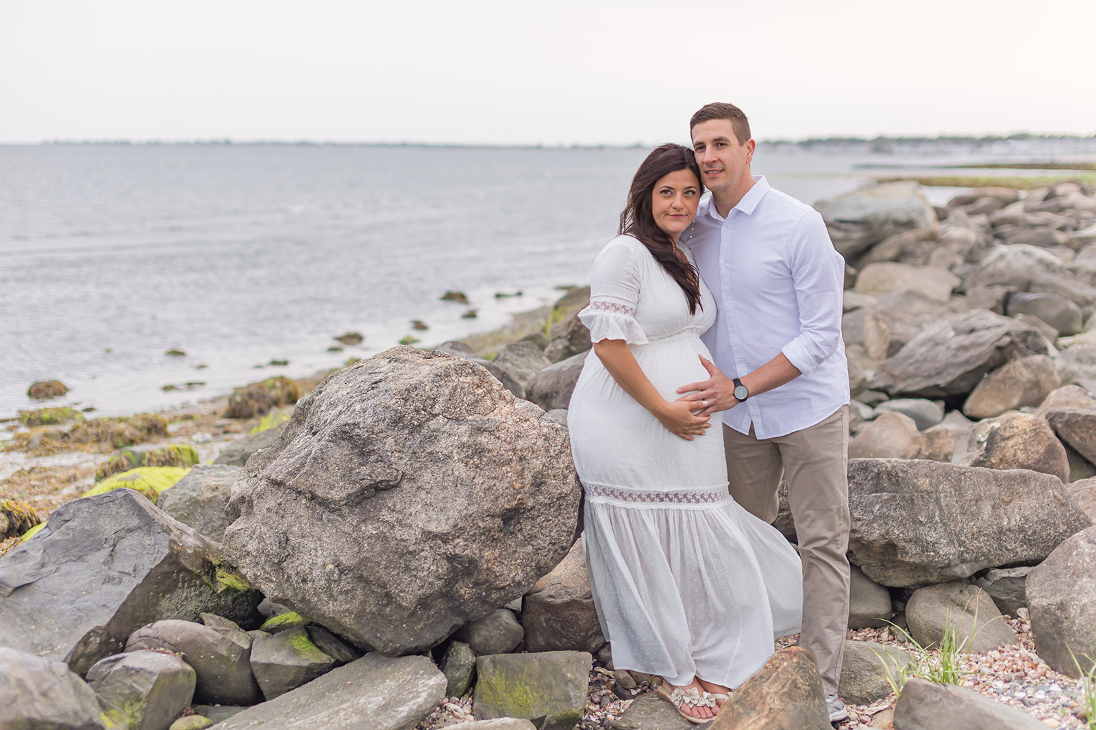 A mom to be in a white maternity gown stands in the rocks on the beach while holding the bump with her husband after visiting St. Vincents Birthing Center