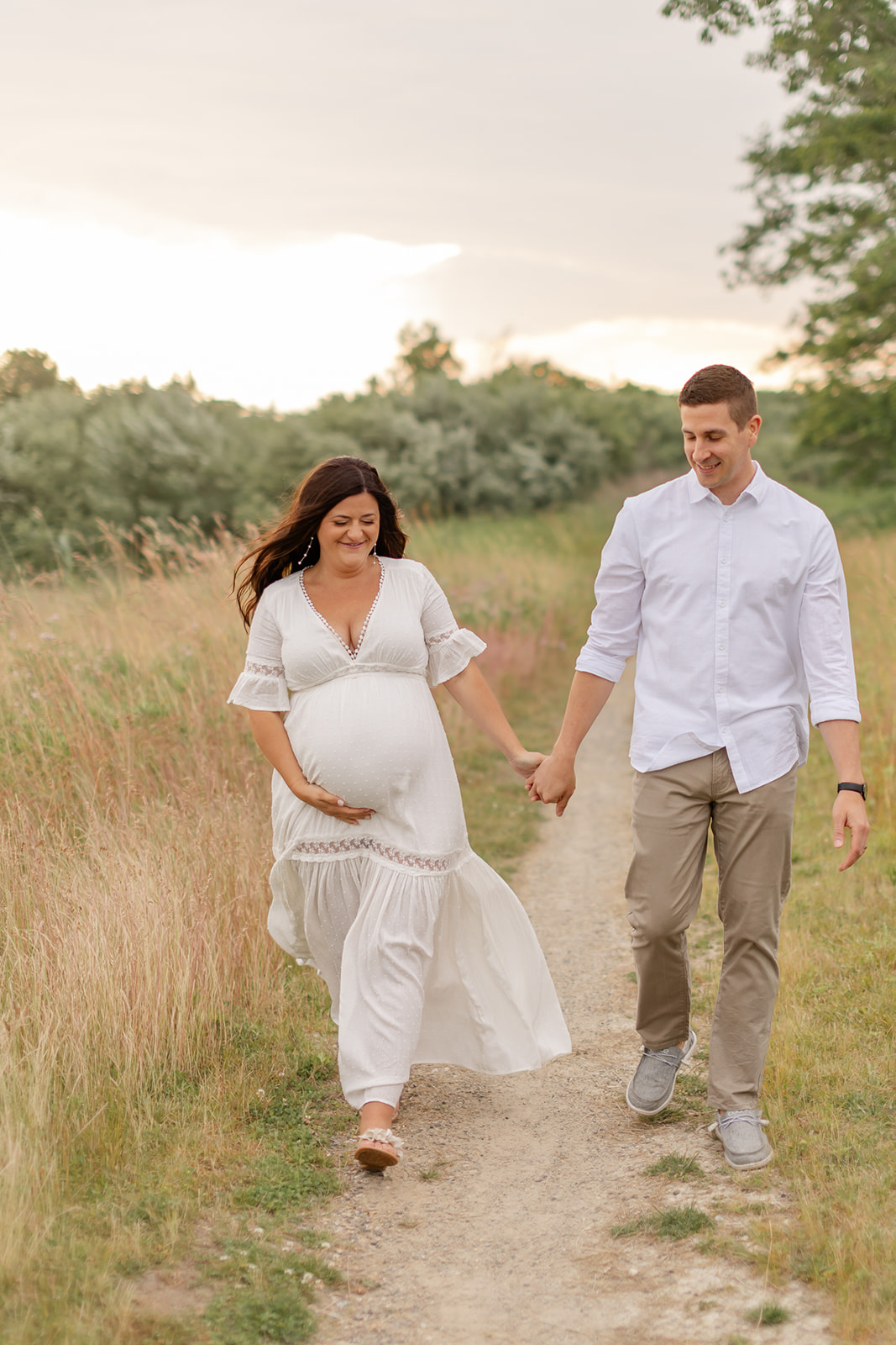 A mom to be holds hands with her husband as they walk down a beach trail at sunset