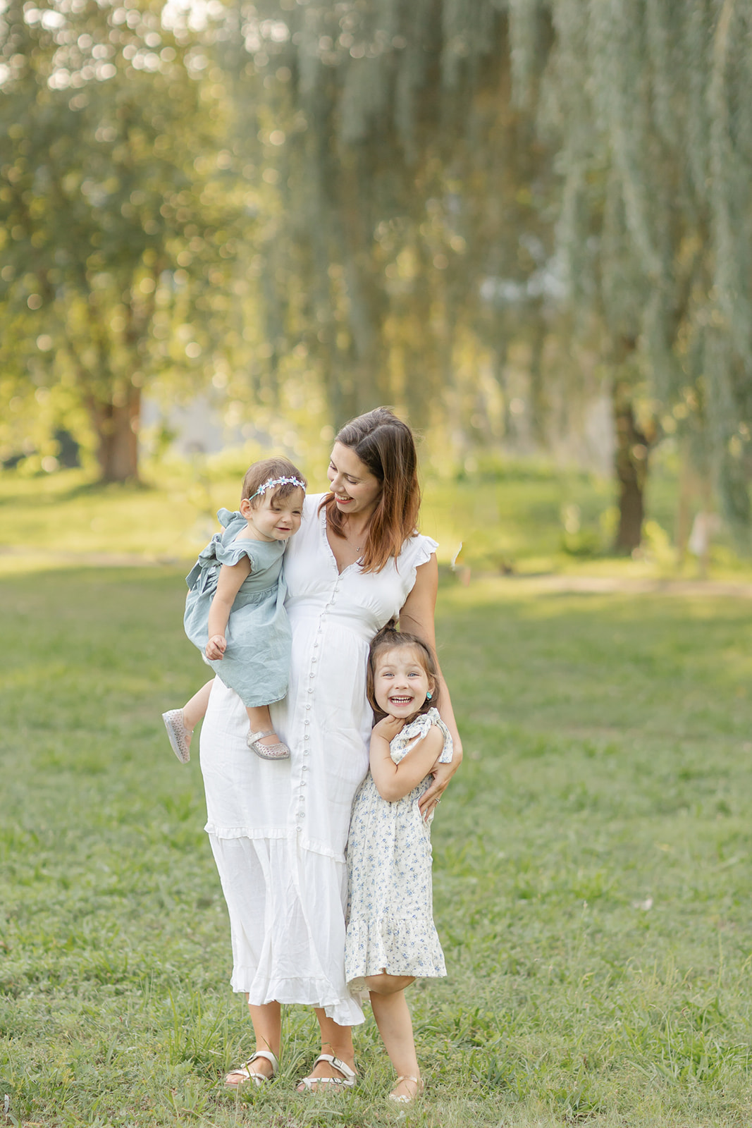 A pregnant mom holds her toddler daughter on her hip while hugging her other daughter at her side in a park