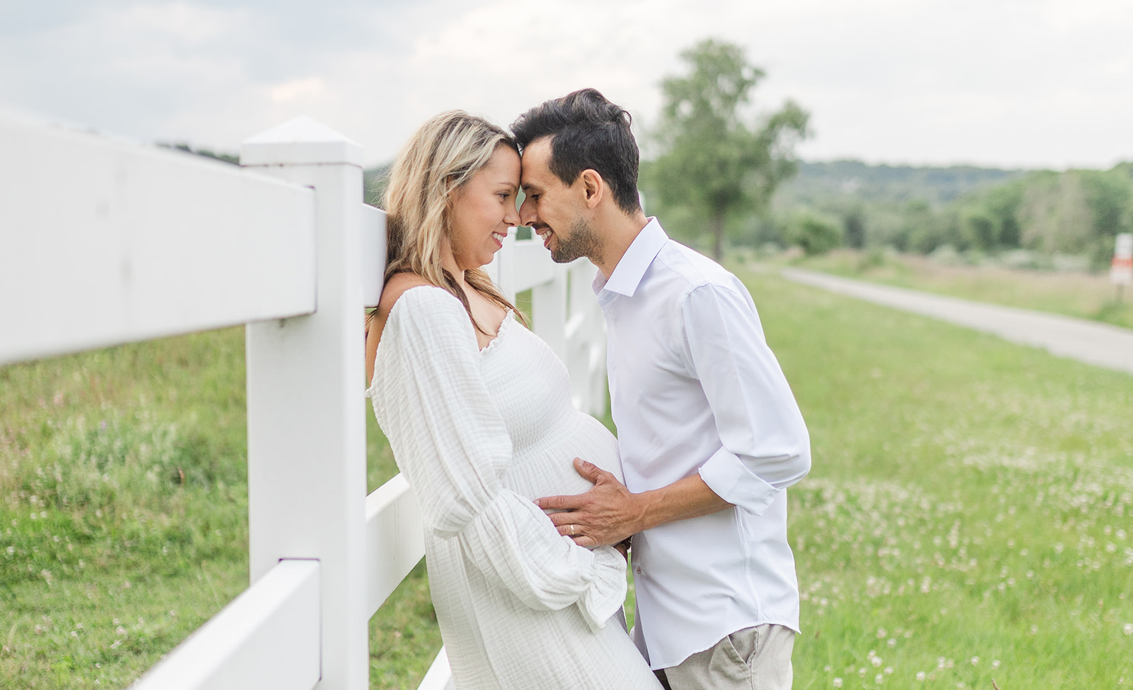 A mom to be leans on a white fence while her and her husband hold the bump in a park