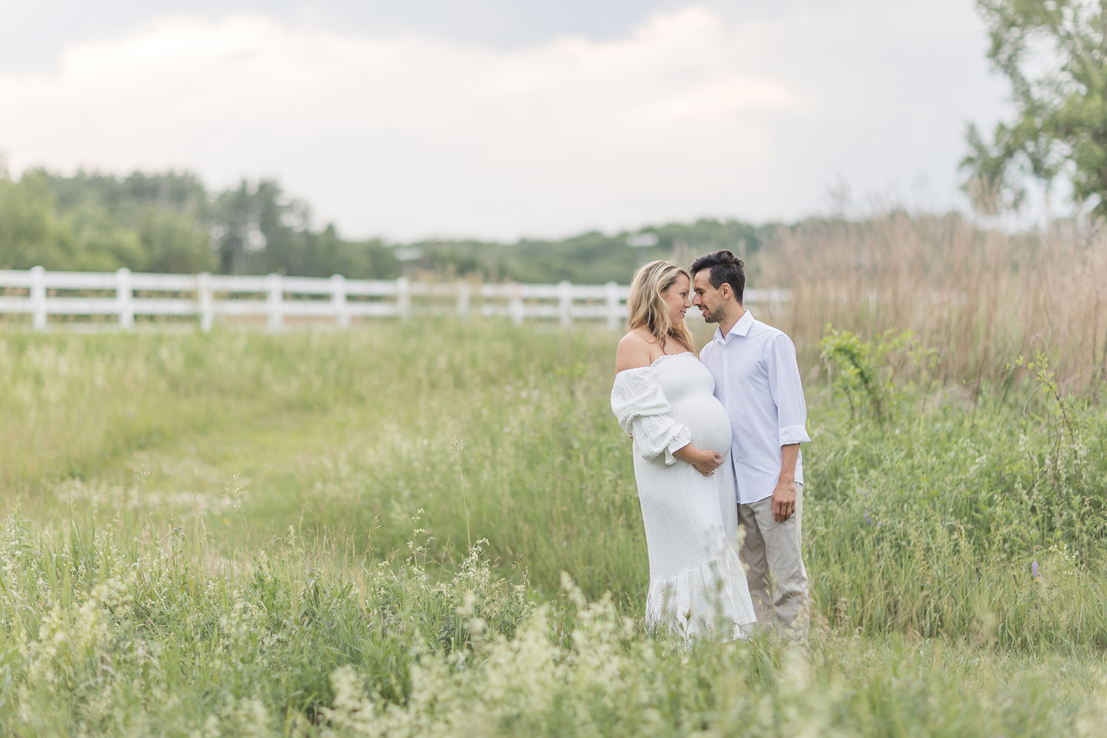 A mom to be in a white maternity gown stands in a park trail about to kiss her husband before visiting Baby shower venues Fairfield CT