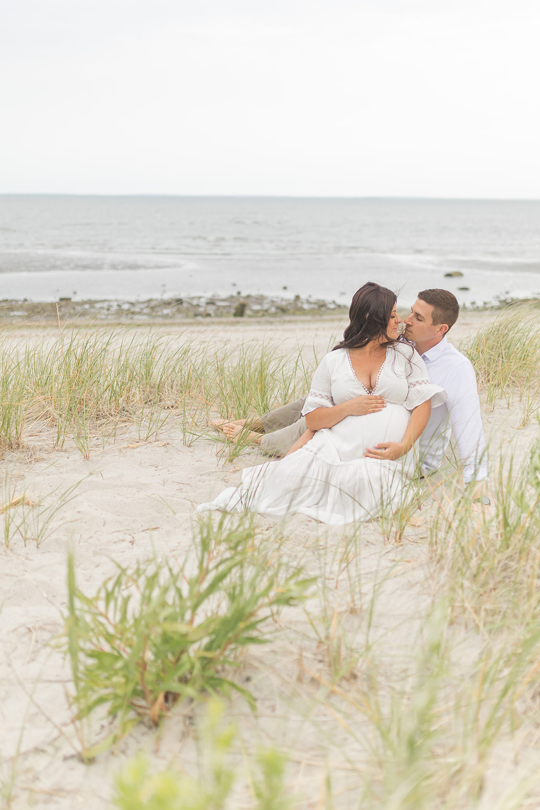 A mom to be sits in the dunes and leans in for a kiss with her husband before visiting Baby Shower Venues Bridgeport CT
