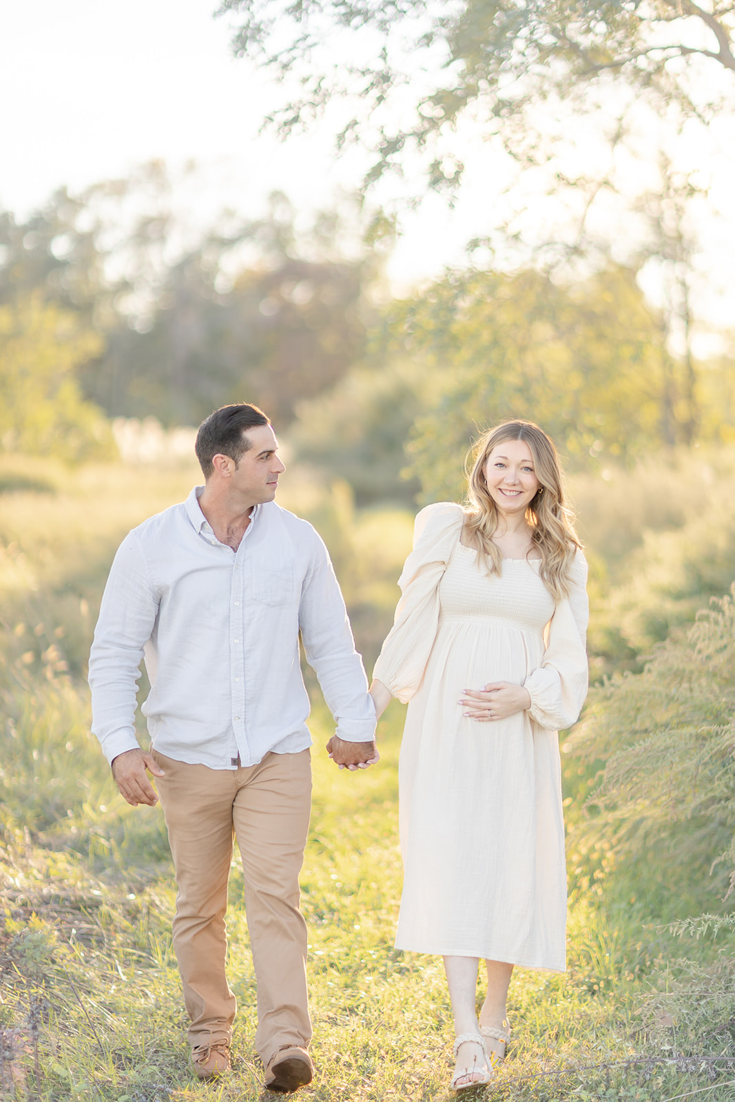 A mom to be walks down a park path at sunset while holding hands with her husband on their babymoon Connecticut