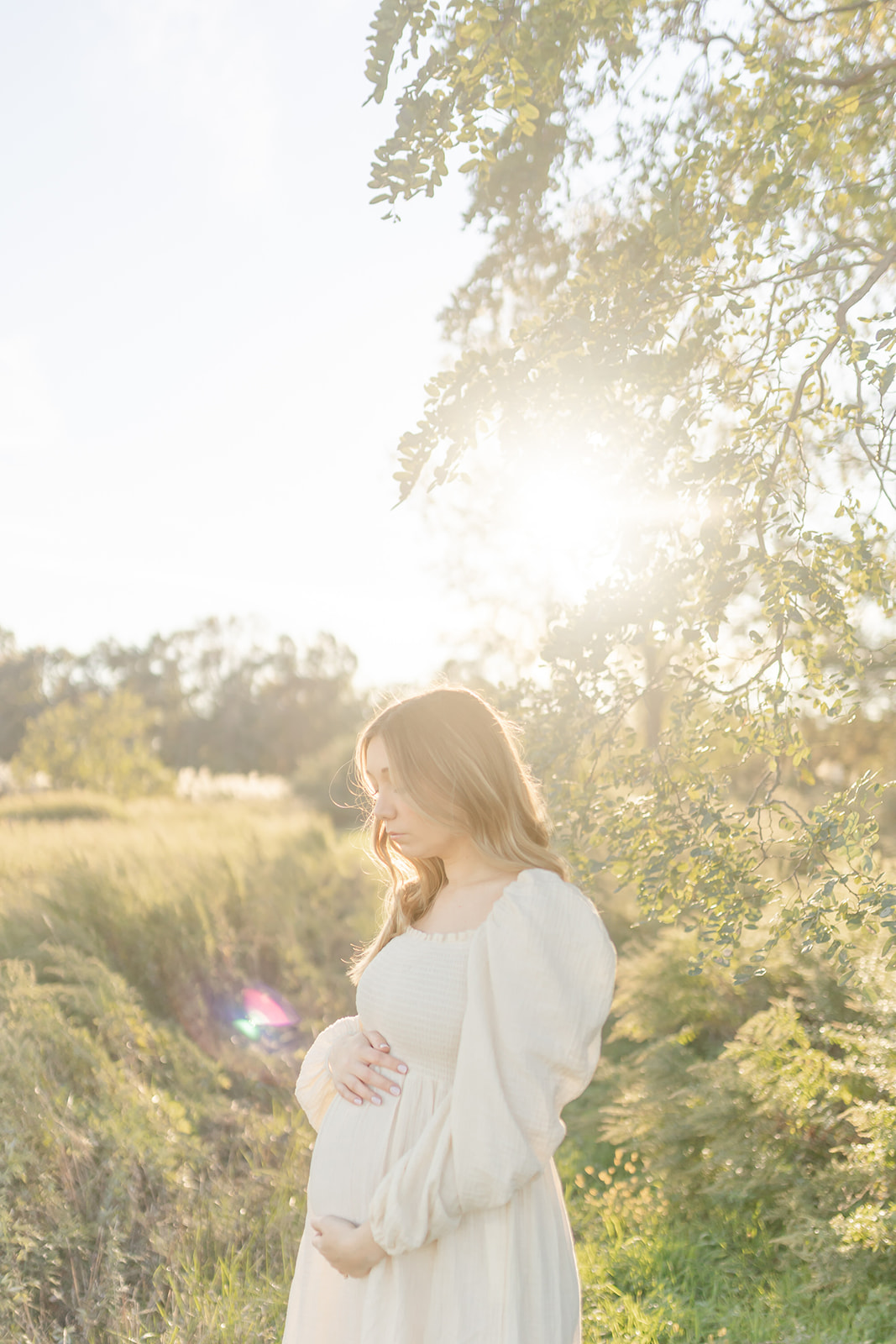 A mom to be holds her bump while standing in a park path at sunset