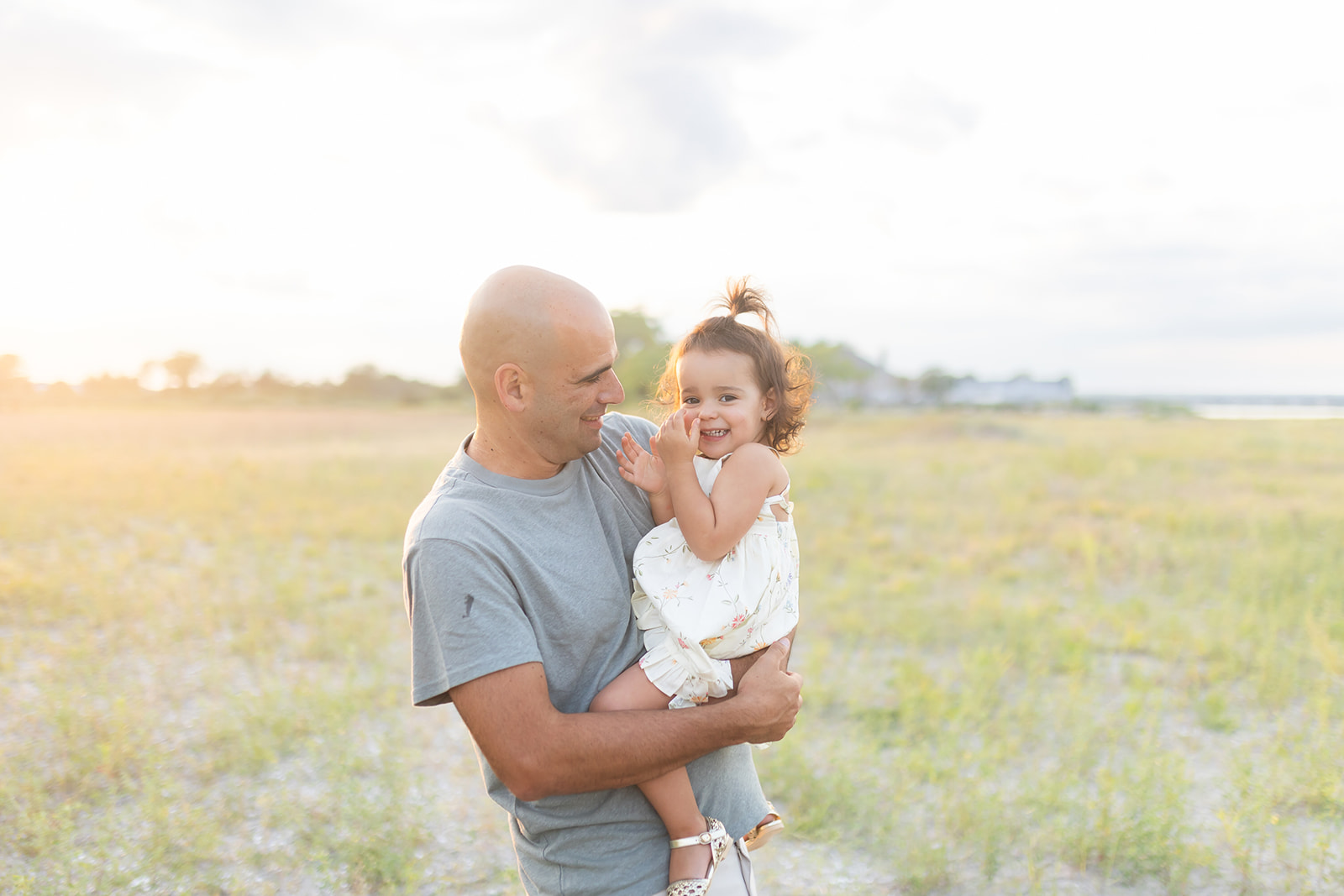 A father in a grey shirt smiles and plays with his toddler daughter on his hip on a beach at sunset