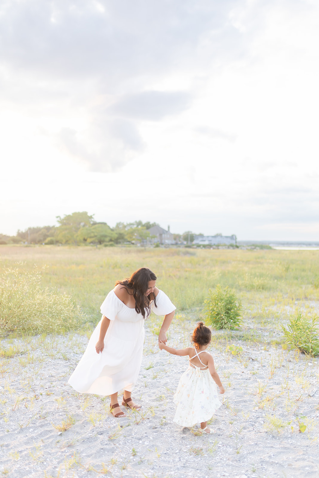 A mother in a white dress dances with her toddler daughter on a beach in a white dress before attending Birthday Parties Fairfield CT