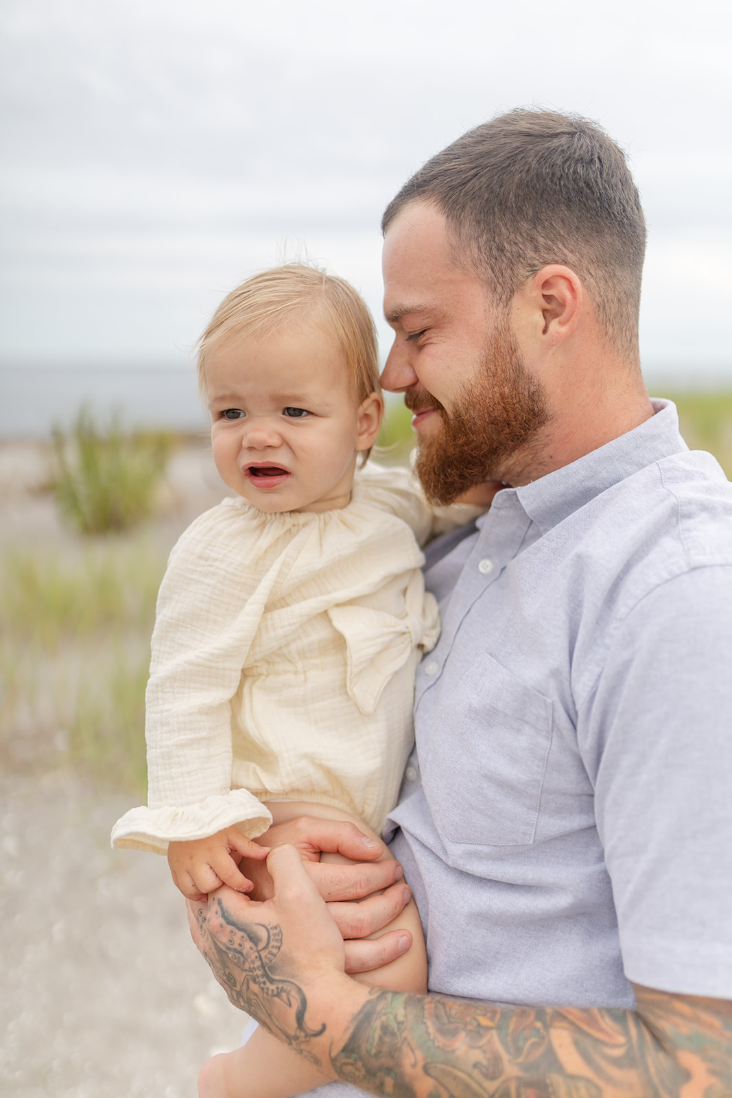 A father stands on a beach holding his toddler daughter in a yellow onesie