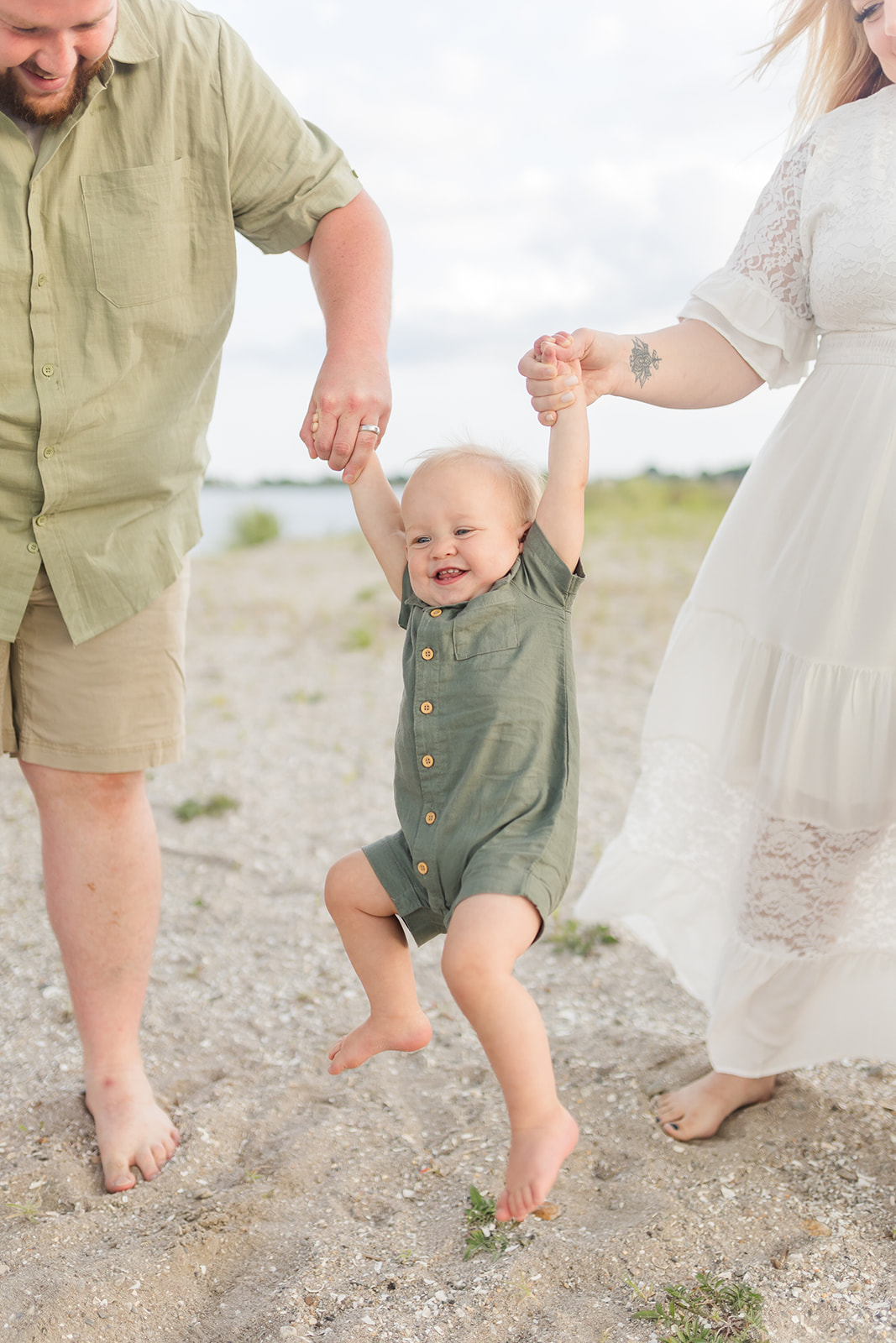 A toddler boy in a green onesie swings in mom and dad's hands on a beach