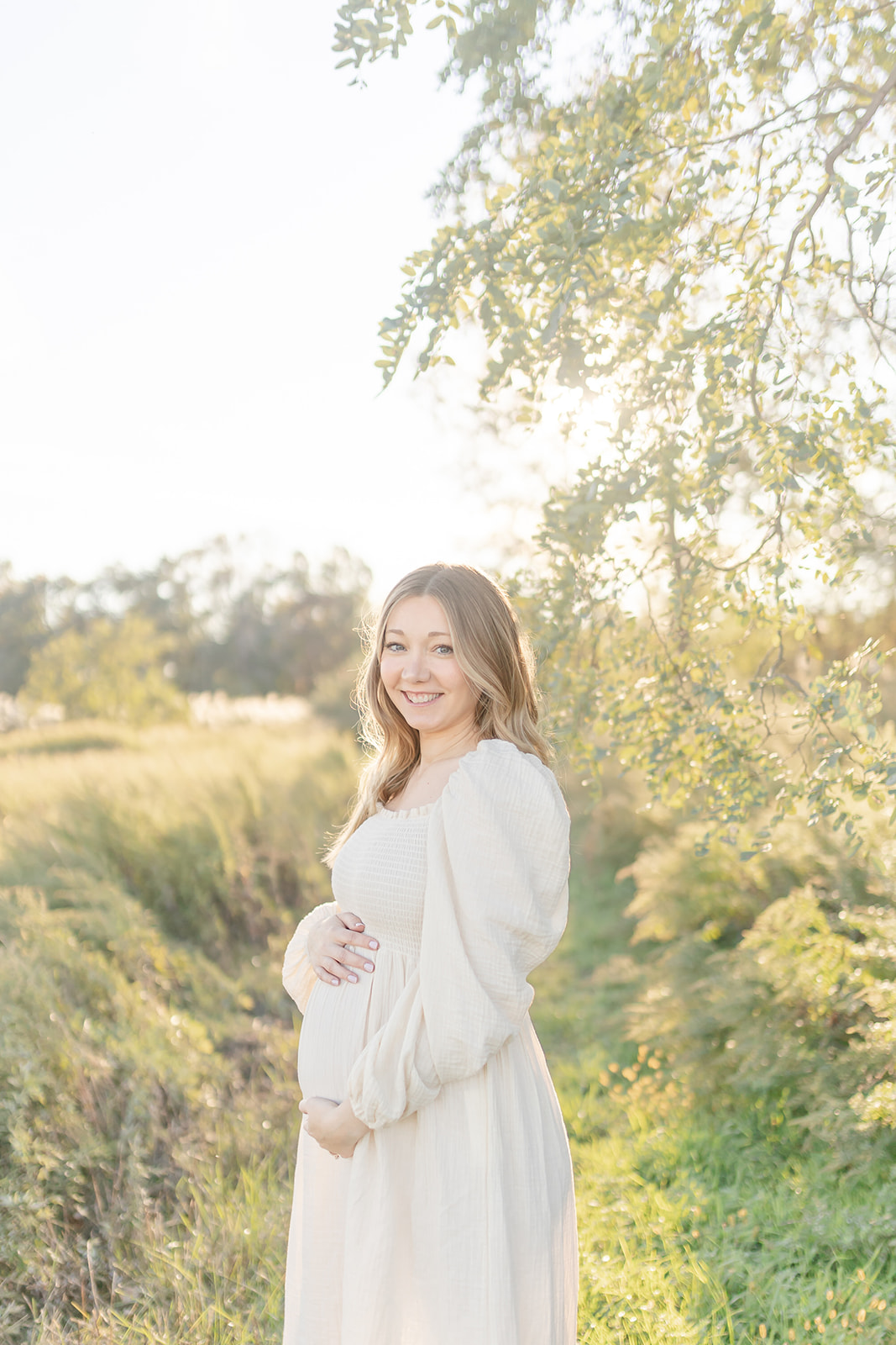 A mom to be stands in a park trail in a white maternity dress at sunset thanks to a Chiropractor in Fairfield CT