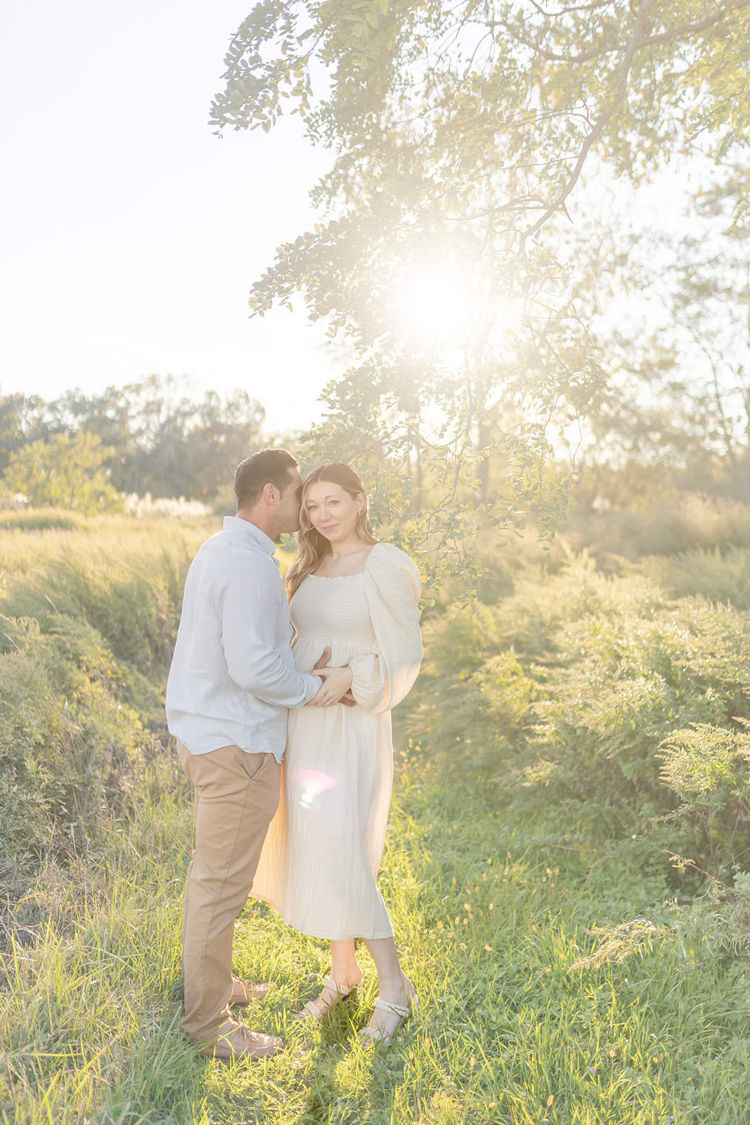 A dad kisses his pregnant wife under a tree at sunset in a park trail after visiting a Chiropractor in Fairfield CT