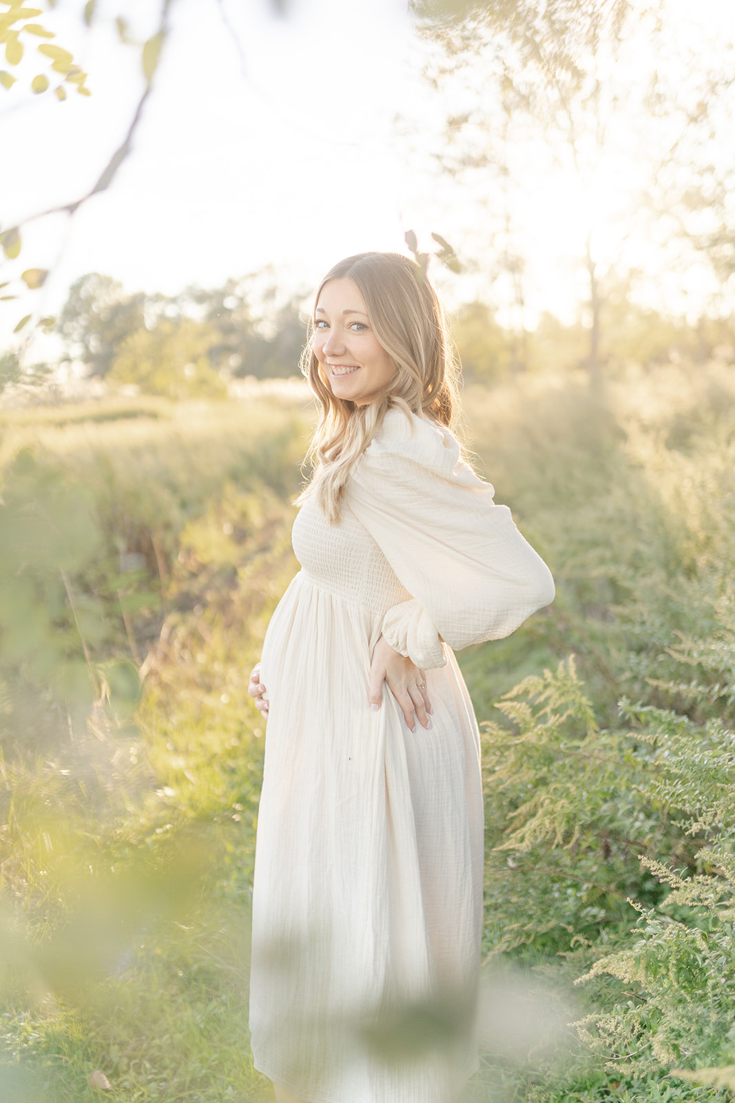 A mother to be rests a hand on her hip while standing in a grassy field at sunset after visiting a CT Birth Center