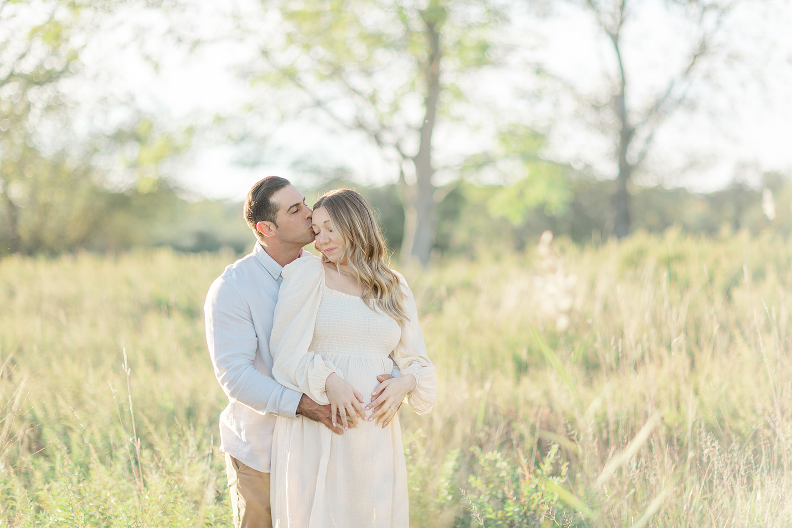 A mom to be is kissed by her husband as her hugs her in a grassy field at sunset after visiting CT Birth Center