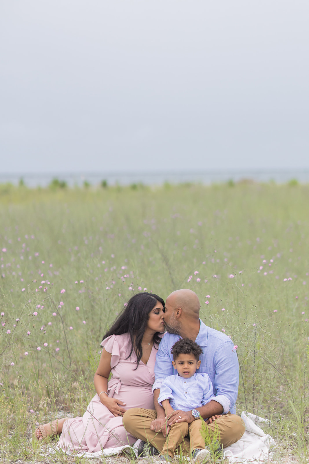 A pregnant mom kisses her husband as they sit on a picnic blanket at the beach with their toddler son in dad's lap after visiting Day Spas Fairfield CT