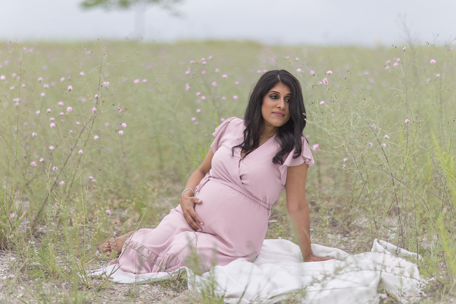 A mom to be in a pink maternity gown sits in a field of wildflowers near a beach on a blanket after visiting Day Spas Fairfield CT