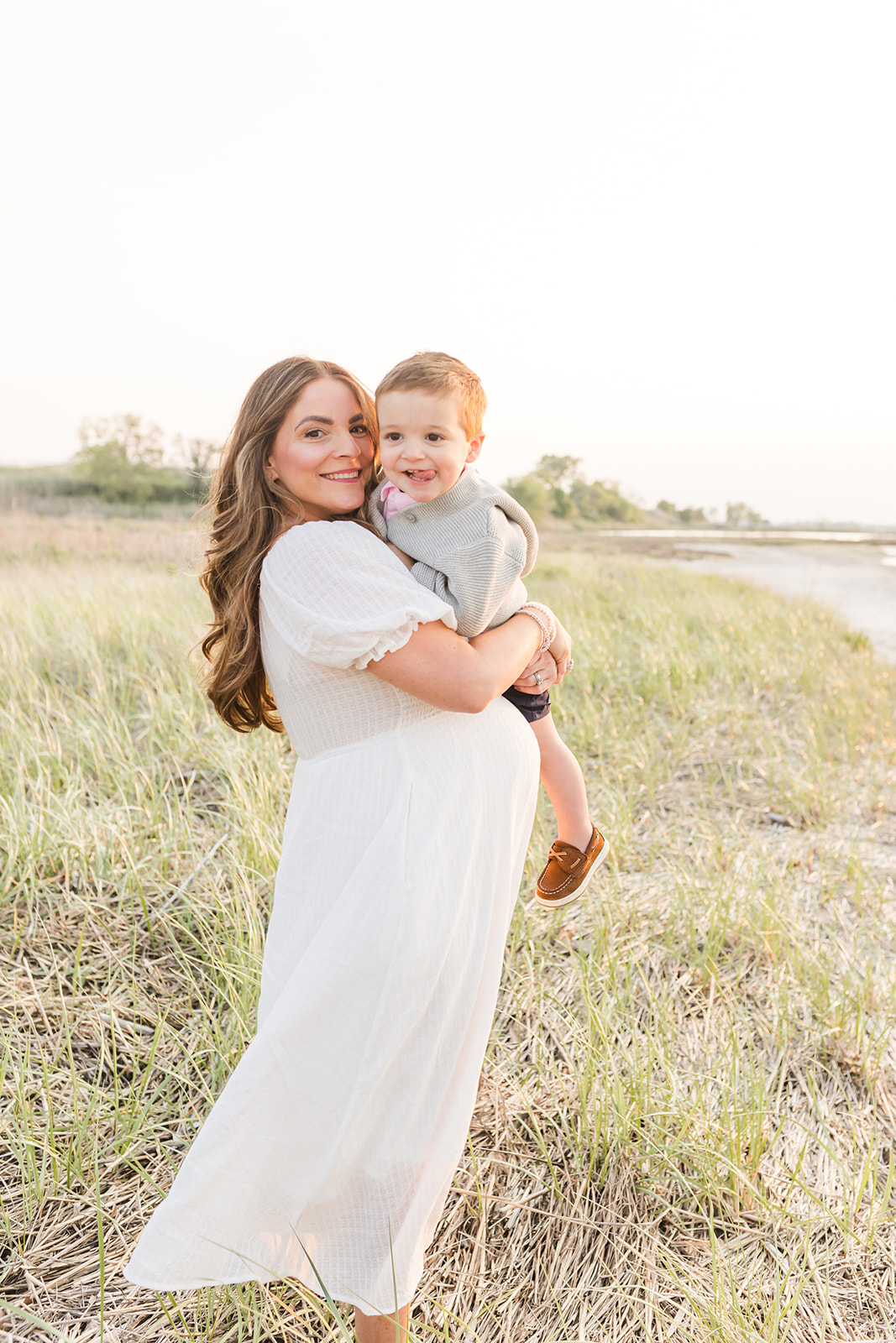 A pregnant mom in a white dress stands on a beach holding her happy toddler son at sunset