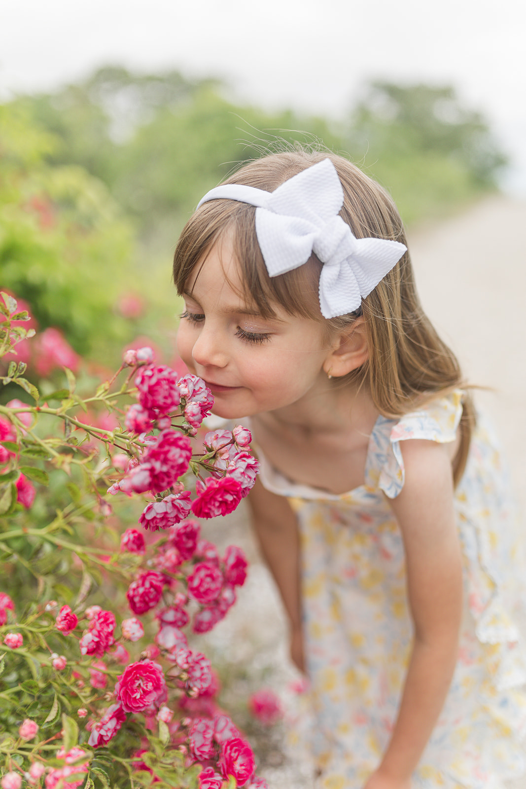 A young girl in a white bow smells some pink wildflowers on a beach trail before attending Fairfield Library Storytime