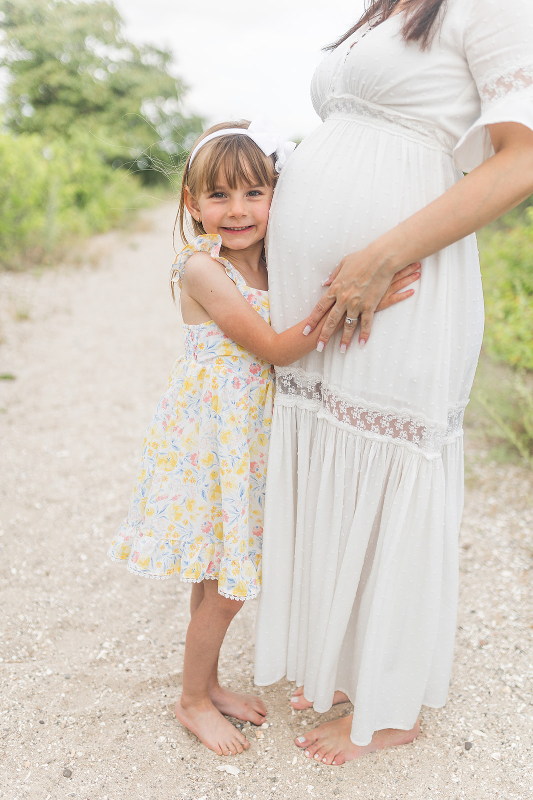 A young girl in a yellow floral dress hugs onto her pregnant mom's bump while standing in a beach path before attending Fairfield Library Storytime